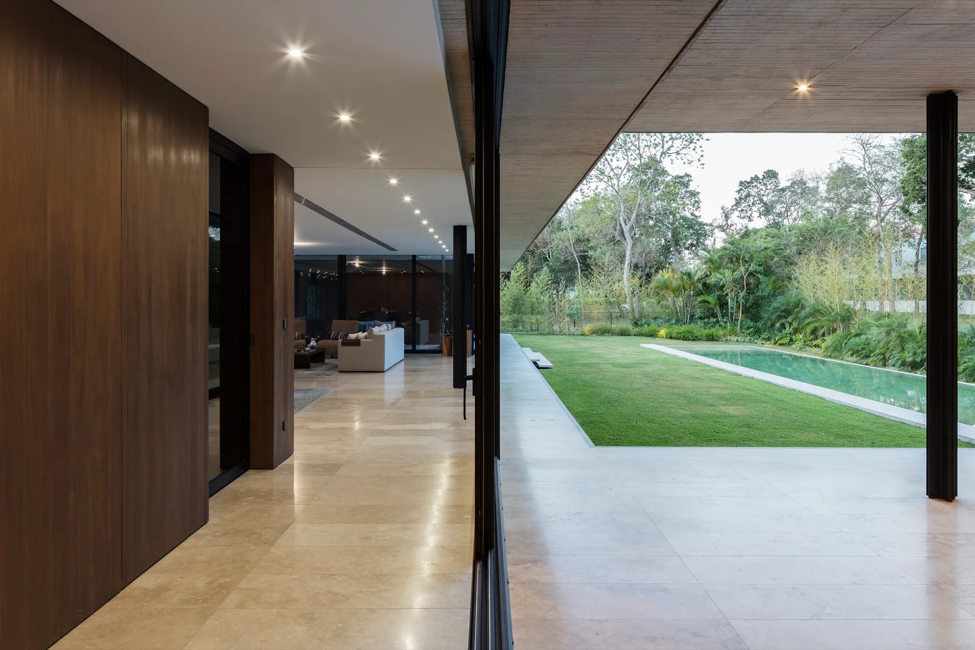 Large sliding glass doors open from a modern living area with beige stone floors onto a covered concrete patio next to a lawn and pool.