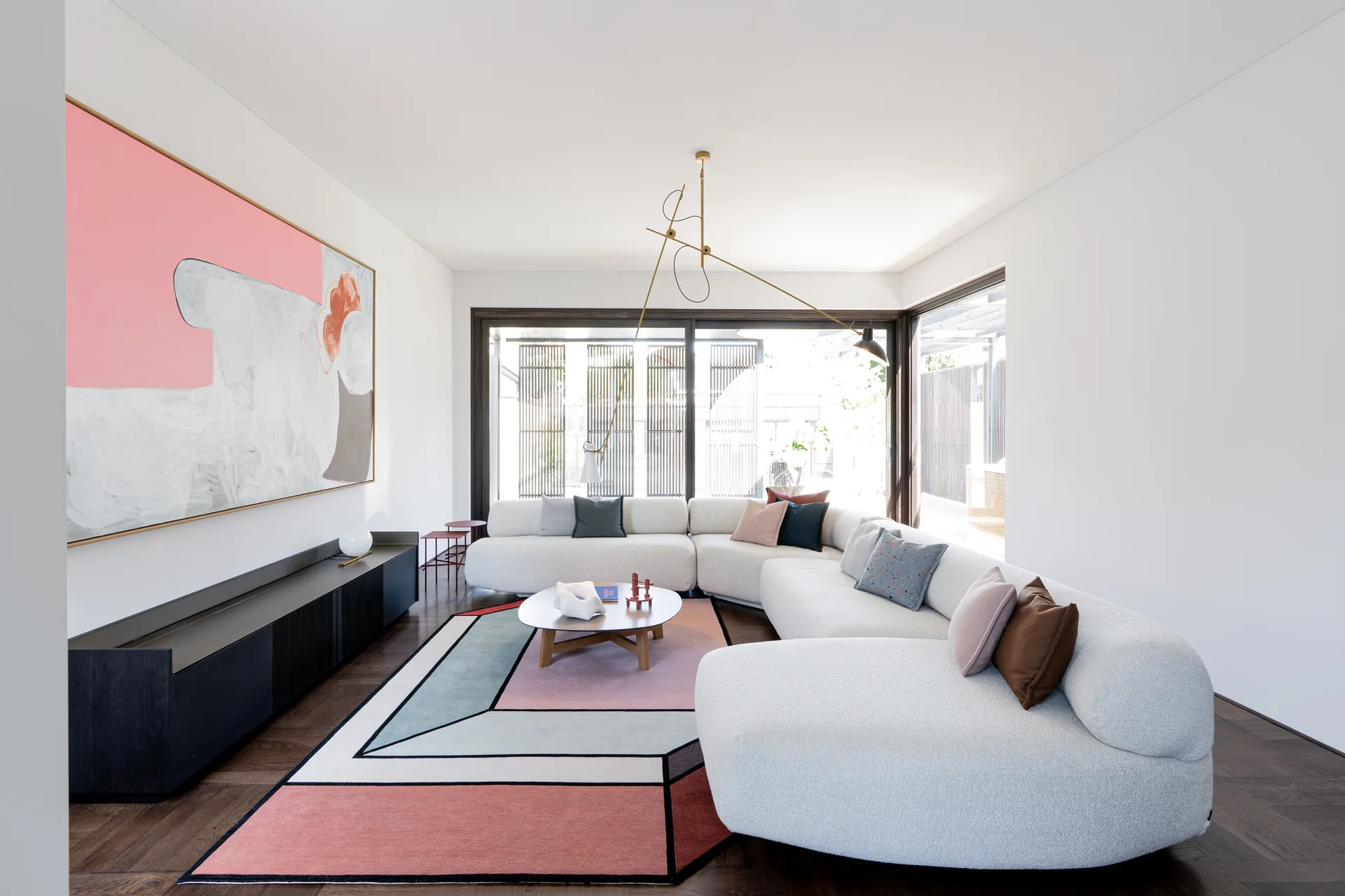 A spacious living room featuring a cream curved sectional sofa, a pink and grey geometric area rug, and a large abstract painting on a white wall.