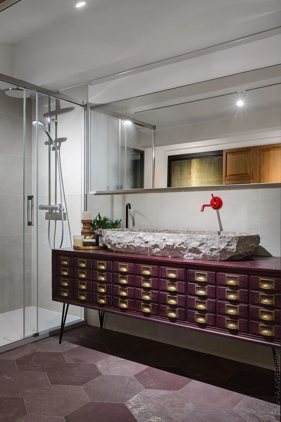 A modern bathroom vanity made from a burgundy apothecary chest with gold pulls, featuring a raw stone vessel sink and a red faucet.