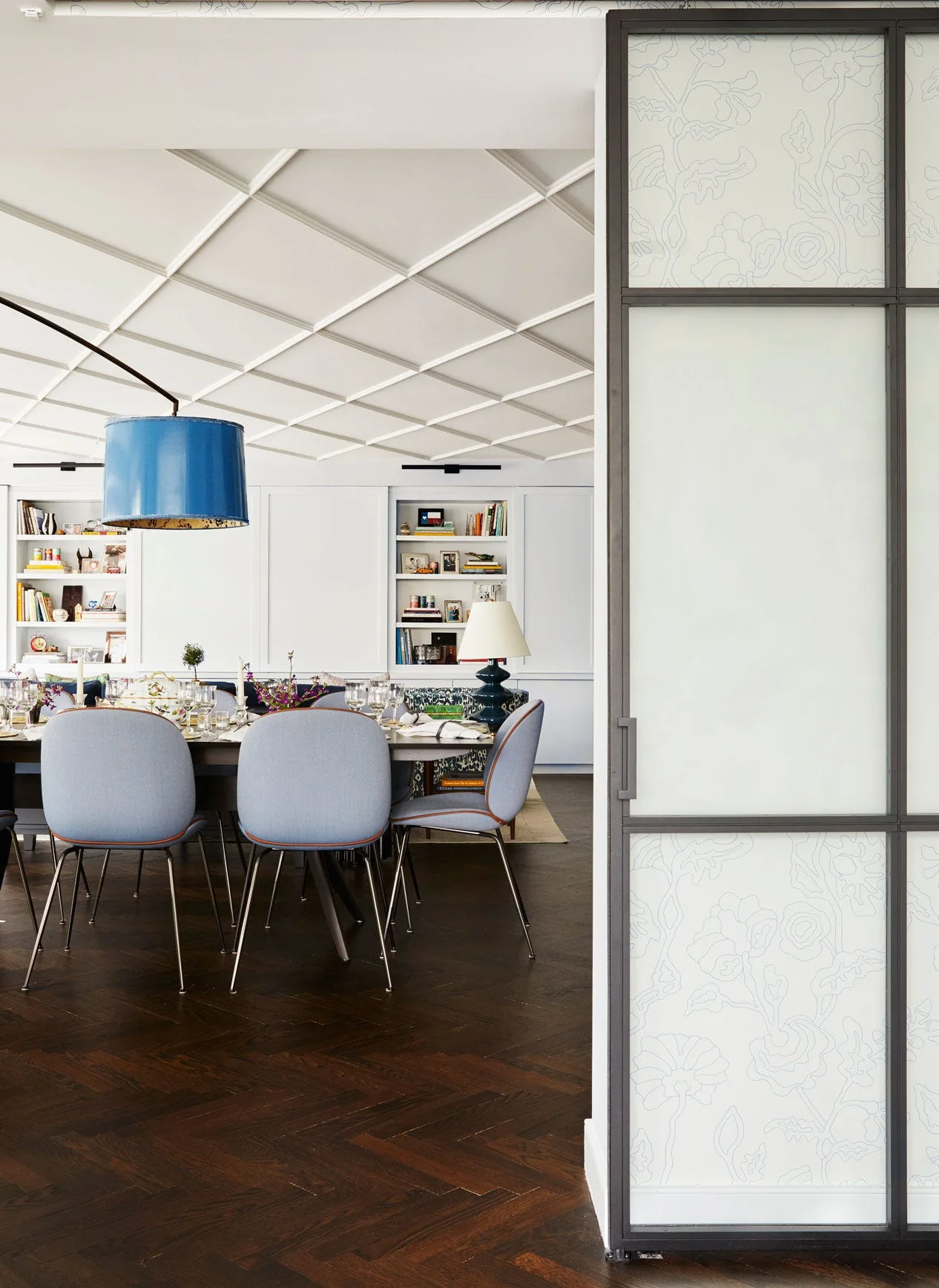A dining room features light blue chairs around a dark table beneath a diagonal grid ceiling, with a dark herringbone wood floor and white built-ins.