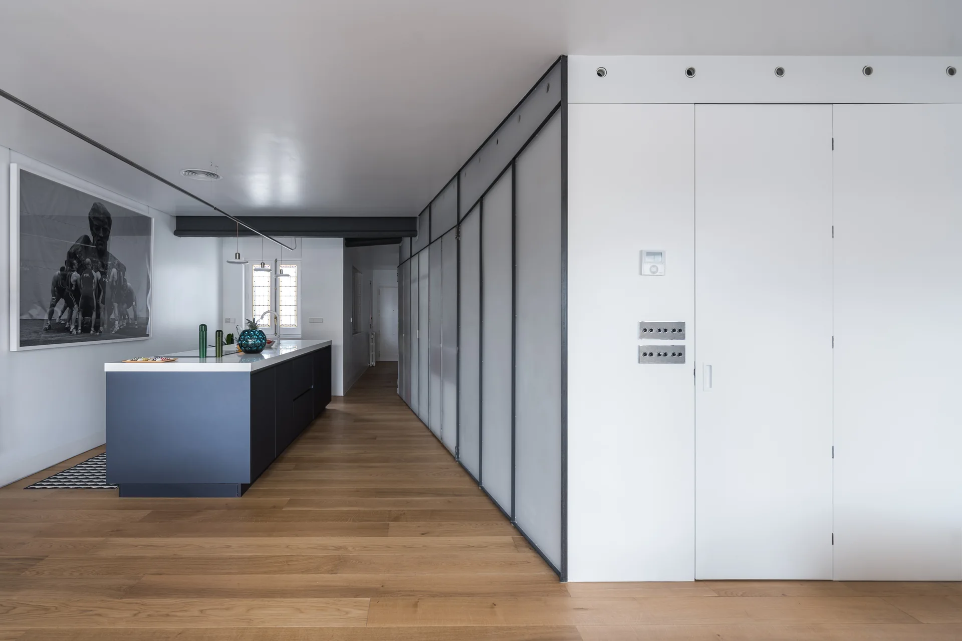 A minimalist kitchen features a dark blue island, light wood flooring, a black-and-white art piece, and a long wall of modular panels.