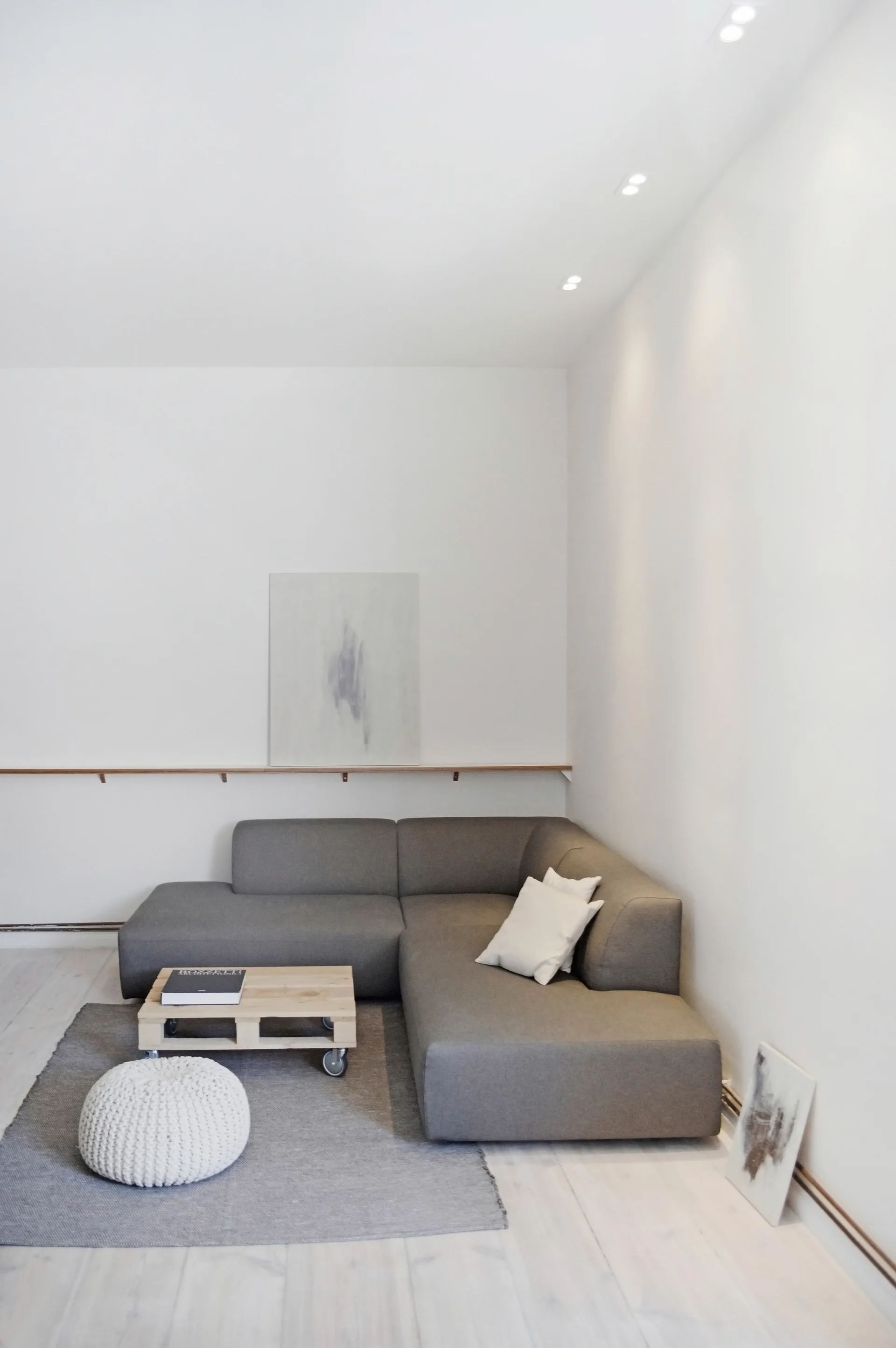 A minimalist living room features a grey sectional sofa, a low wooden pallet coffee table on wheels, a white knitted pouffe, and a thin wood wall ledge.
