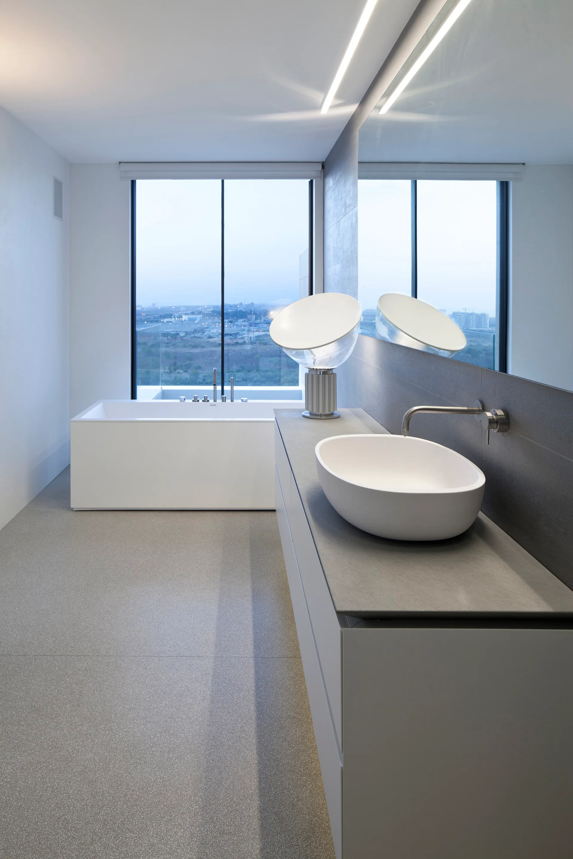 A modern grey bathroom featuring a long vanity with an oval vessel sink, a large mirror, and a white rectangular soaking tub beside a large window.