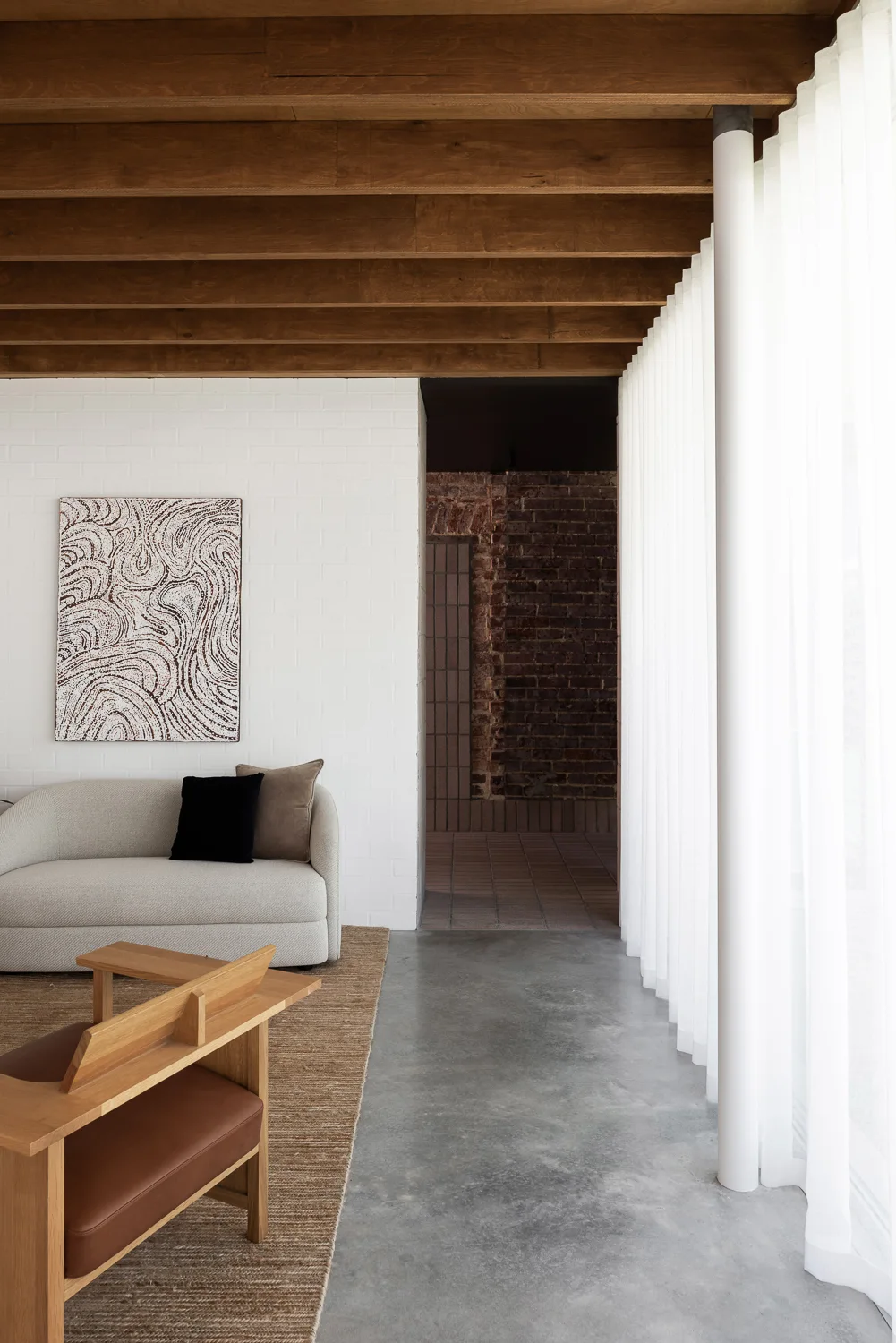 A modern living room features exposed wooden ceiling beams, a white painted brick wall with textured art, a light grey sofa, and concrete flooring.