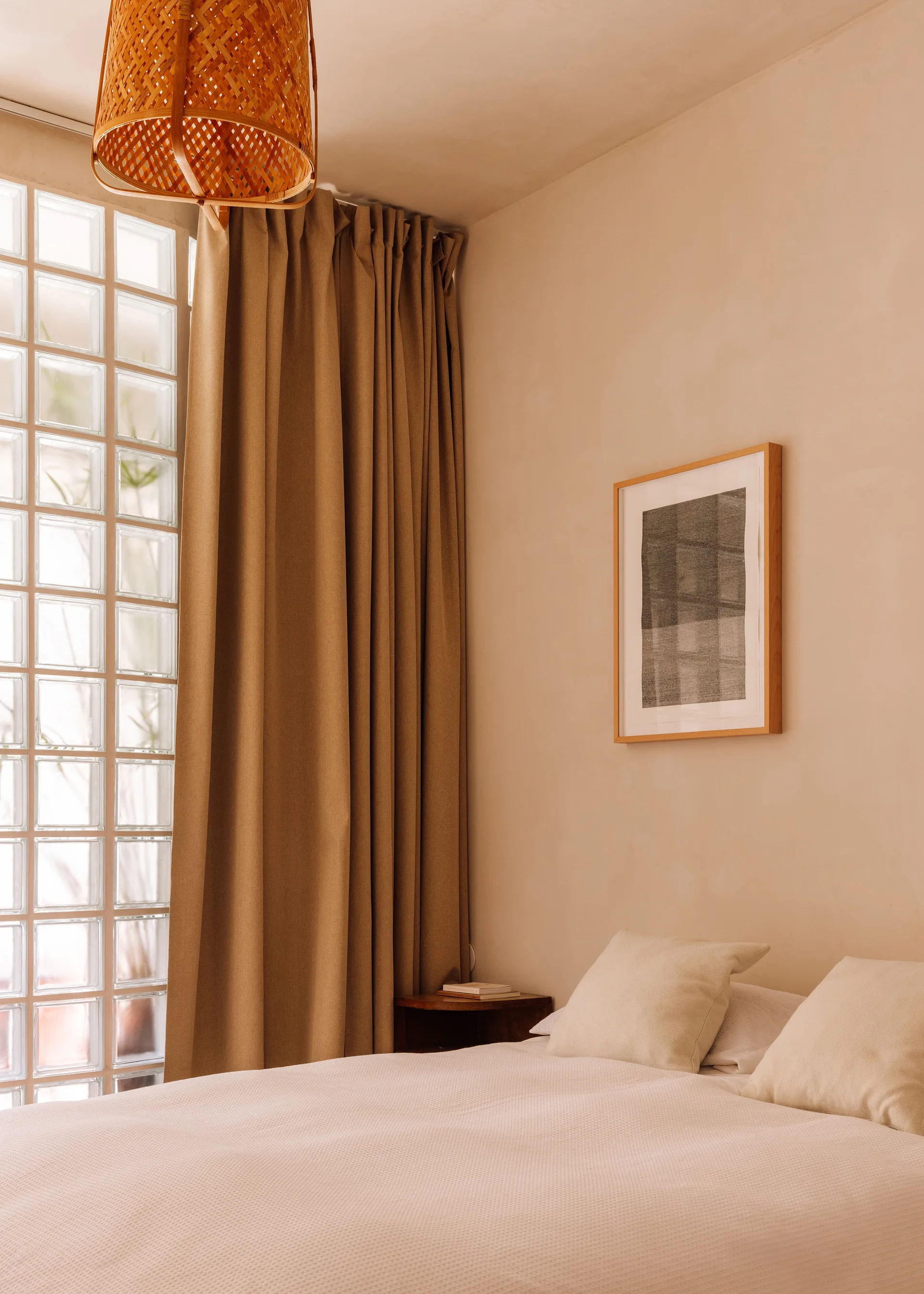 A minimalist bedroom featuring a bed with neutral pillows, a glass block window panel, beige curtains, and a woven wicker pendant light.