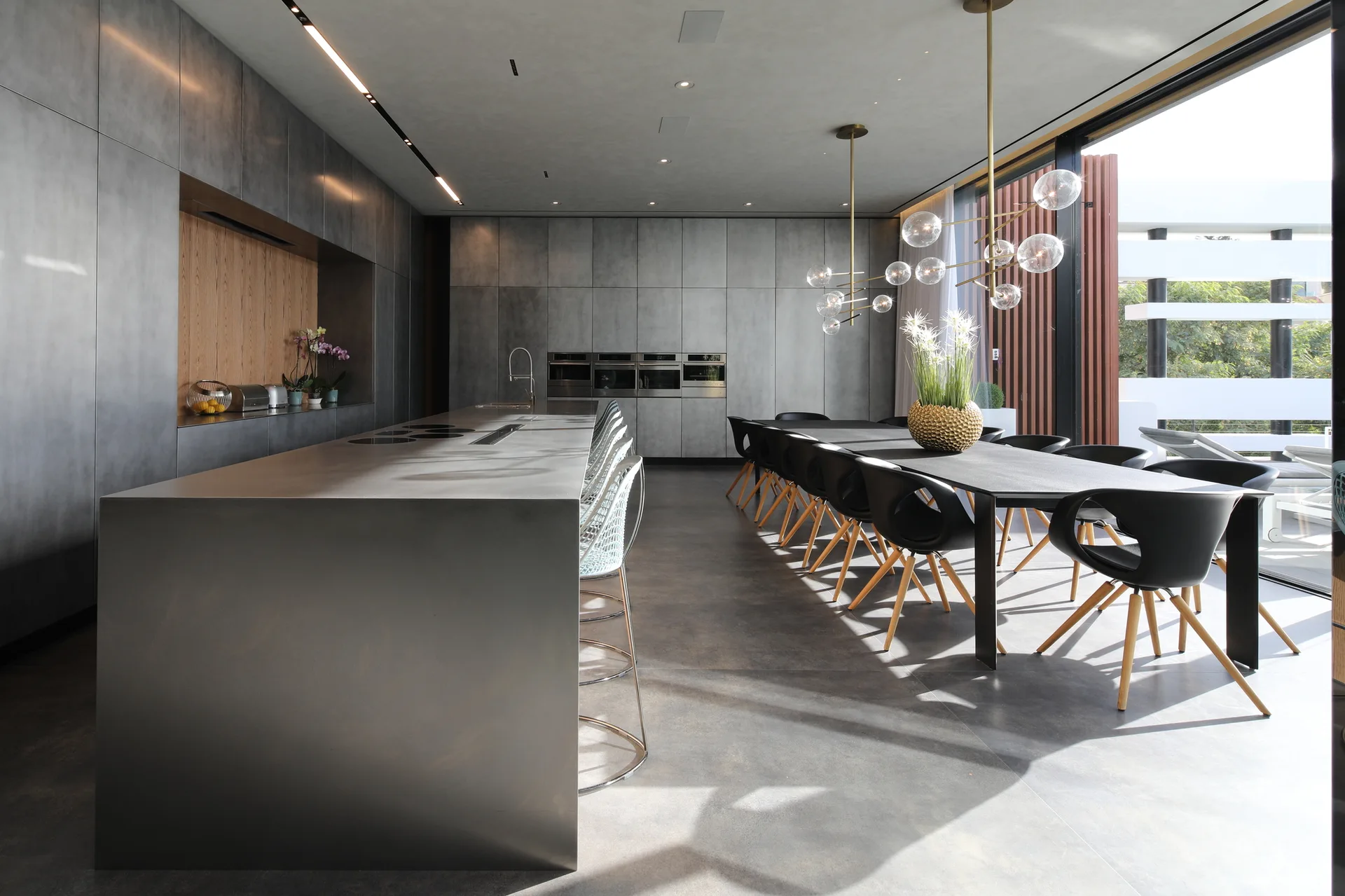 A large gray kitchen features a metal island, matching wall cabinets with a wood niche, and a long dining table under bubble pendant lights.