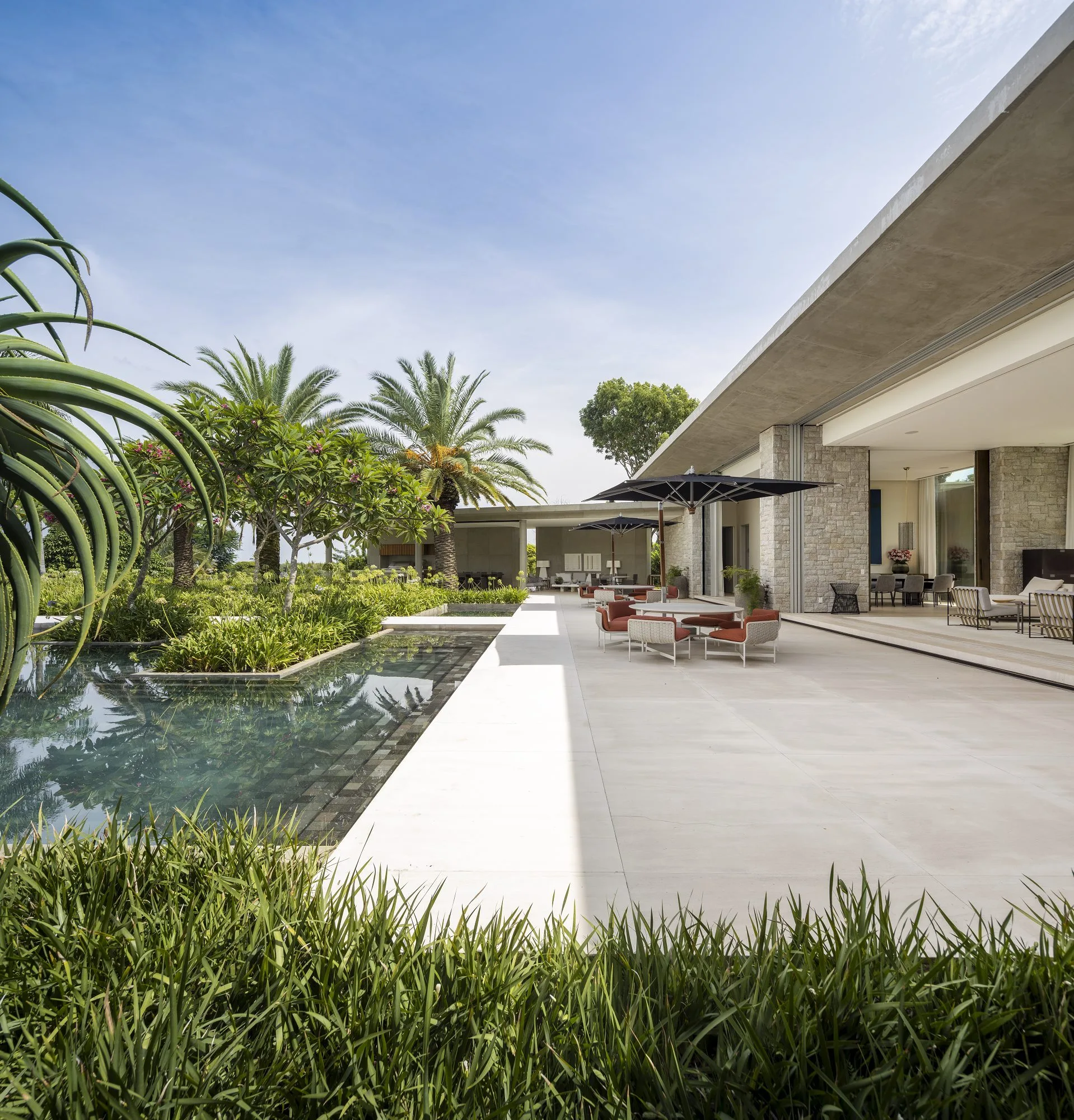 A modern concrete patio features stone-clad walls, poolside dining furniture with black umbrellas, and a swimming pool bordered by tropical plants.