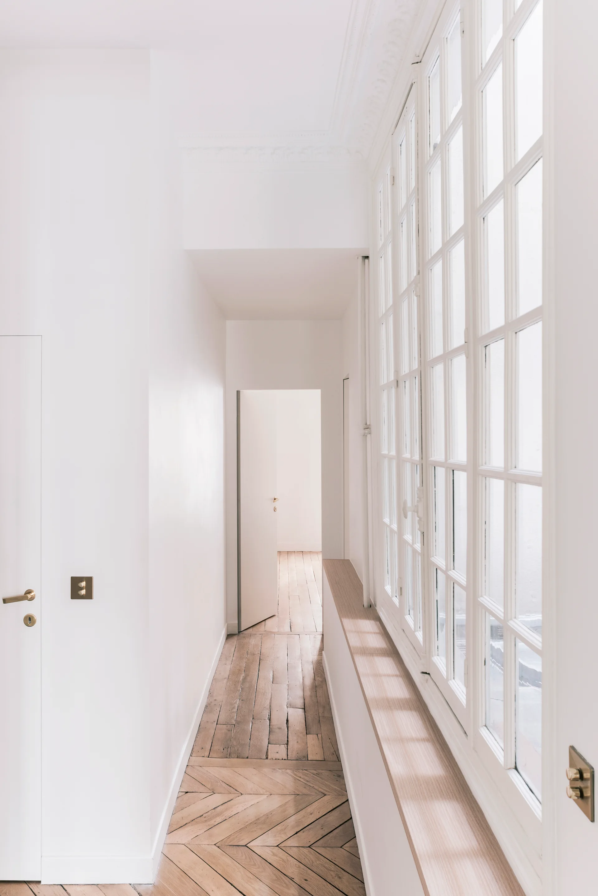 A bright white hallway features herringbone parquet flooring, a long wooden window sill, and a row of tall mullioned windows.