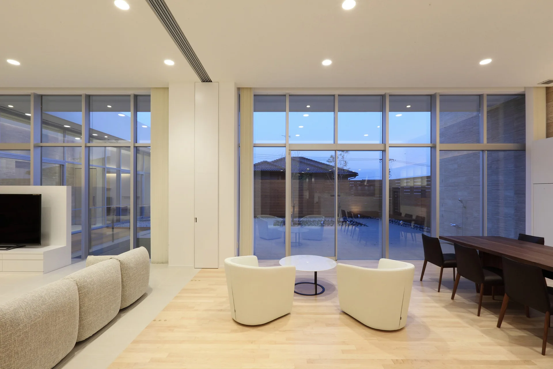 A spacious living and dining area features light wood floors, curved white armchairs, and a large glass facade opening to an outdoor courtyard.