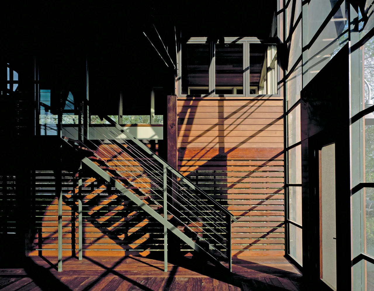 A modern staircase with dark metal cable railings rises against a horizontal slatted wood wall, illuminated by geometric shadow patterns from a nearby window.