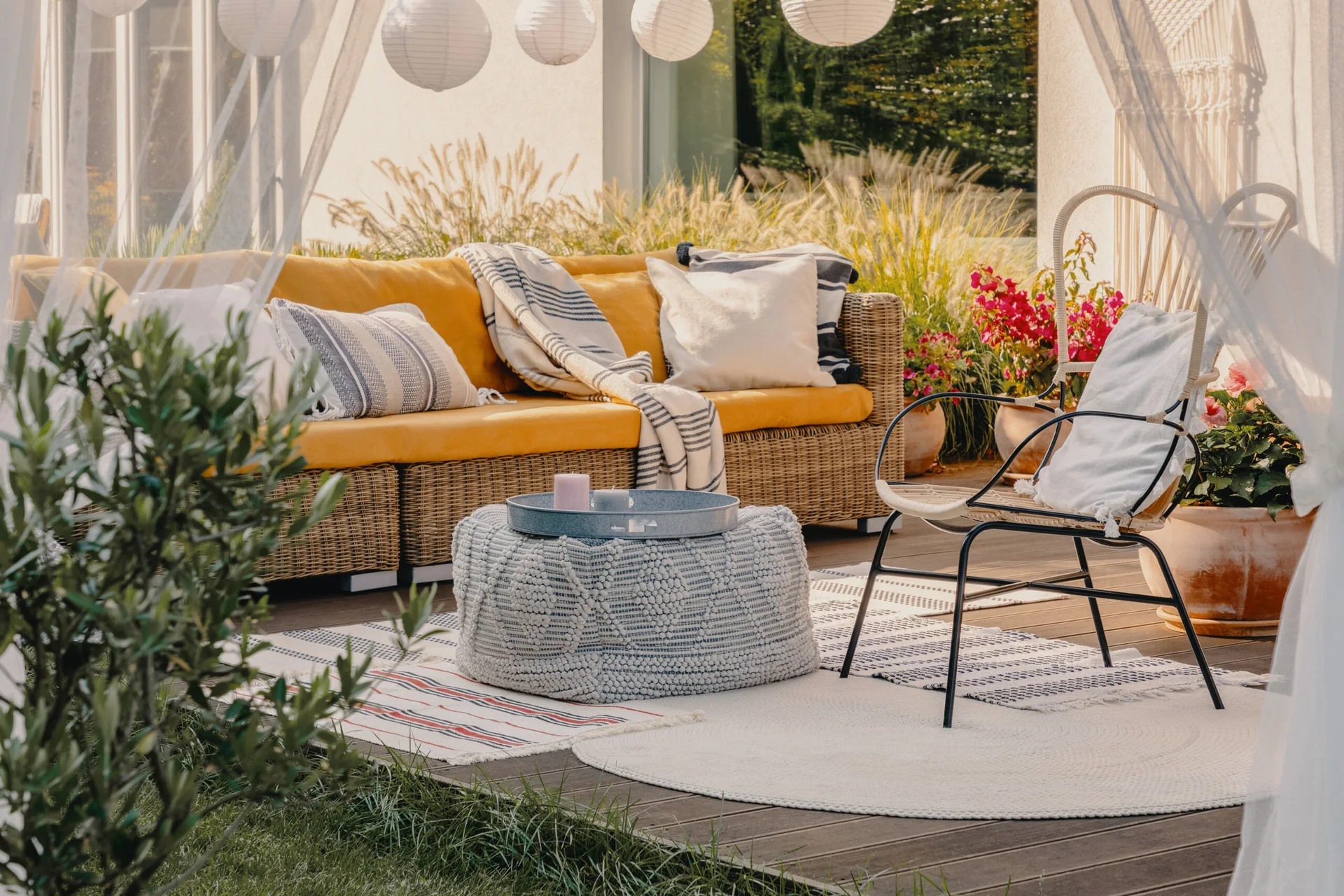 A wooden patio deck features a wicker sofa with bright yellow cushions, a textured grey pouf, layered rugs, and hanging paper lanterns.