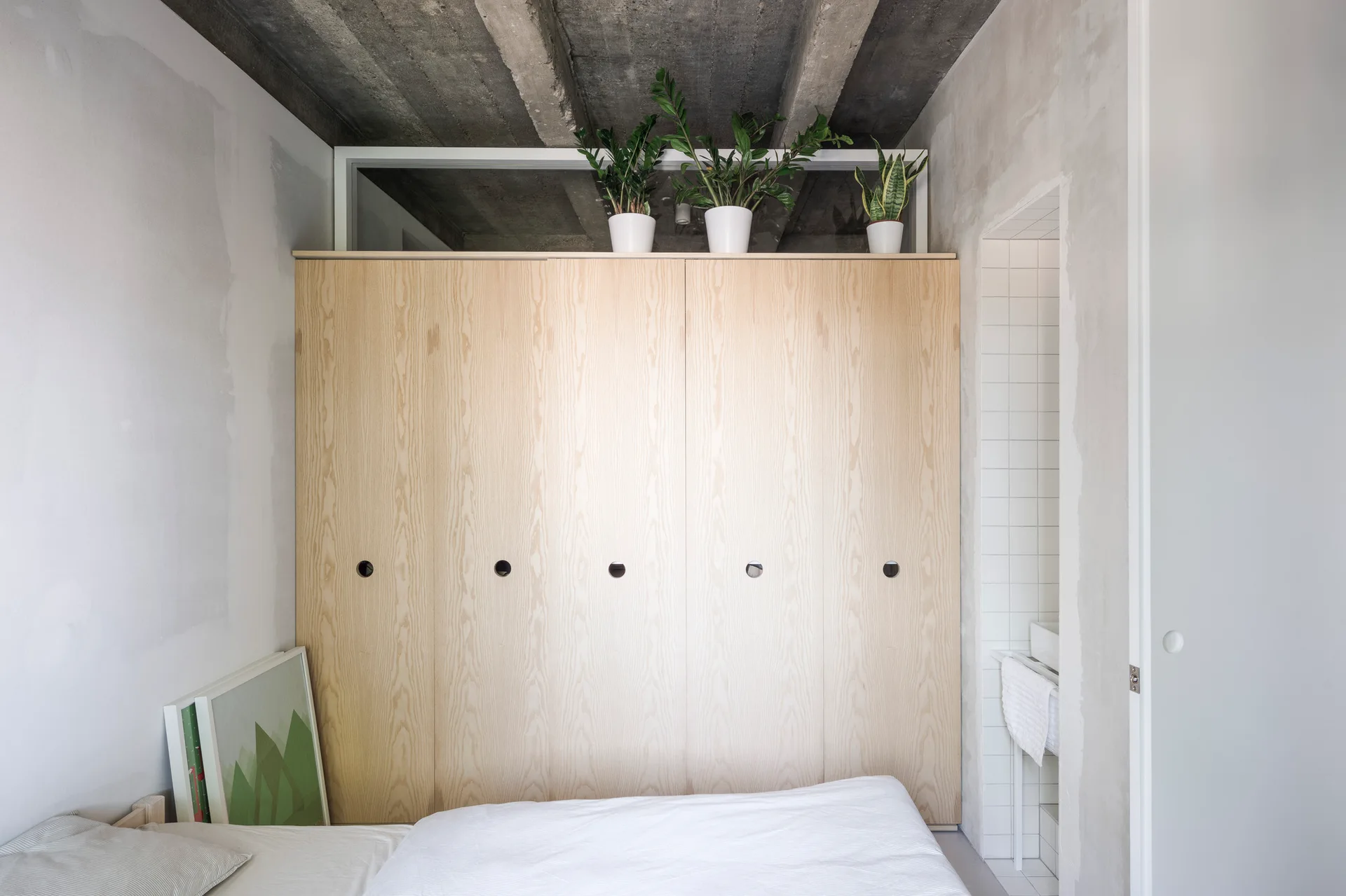 A minimalist bedroom features a large blonde wood wardrobe with circular cutout handles beneath an exposed concrete ceiling.