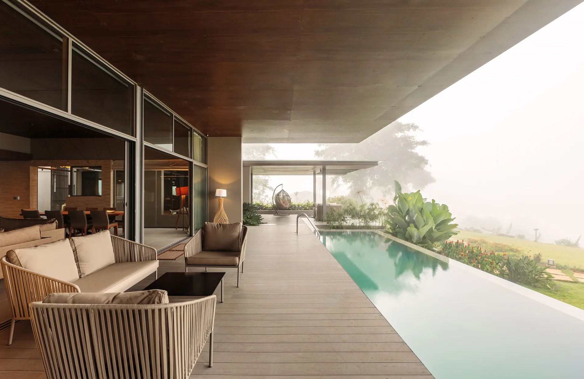A covered wooden patio features a beige woven sofa and chairs next to a long infinity pool overlooking a foggy landscape.