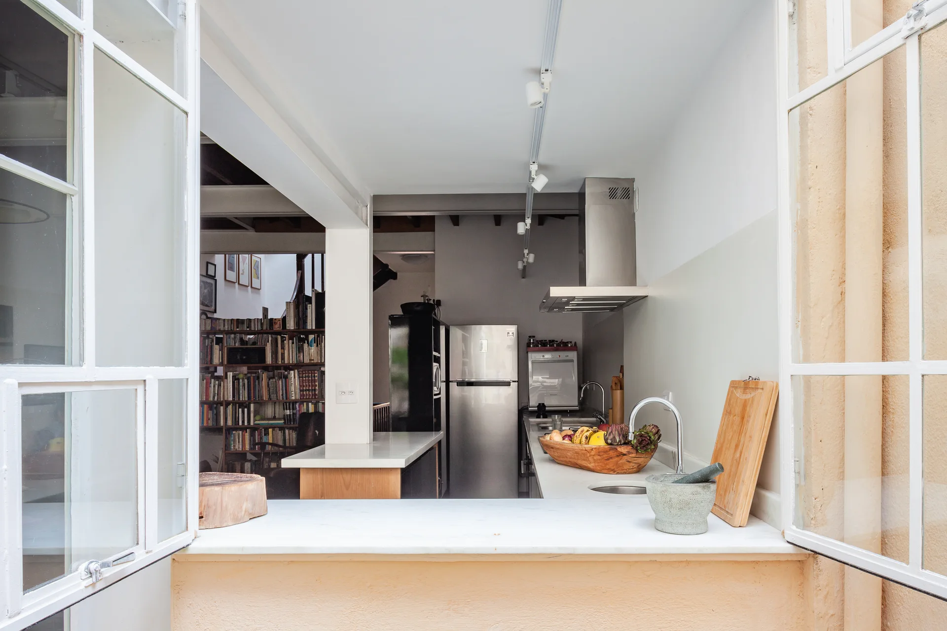 A view through an open white window reveals a bright kitchen with a marble countertop, stainless steel refrigerator, and a bookshelf in the background.