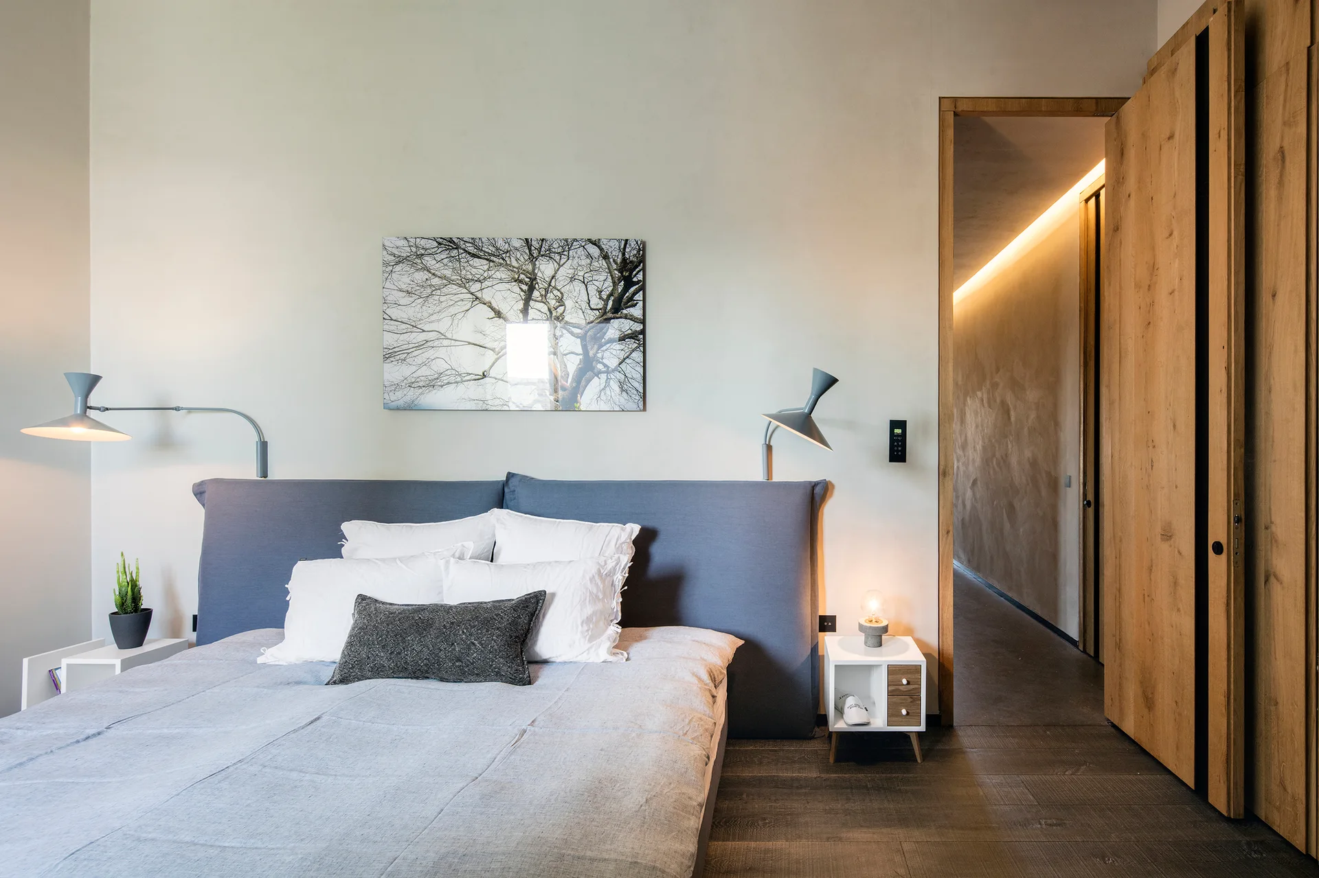 A minimalist bedroom featuring a bed with a blue upholstered headboard, a wood door opening to a hallway, and wall-mounted lamps.