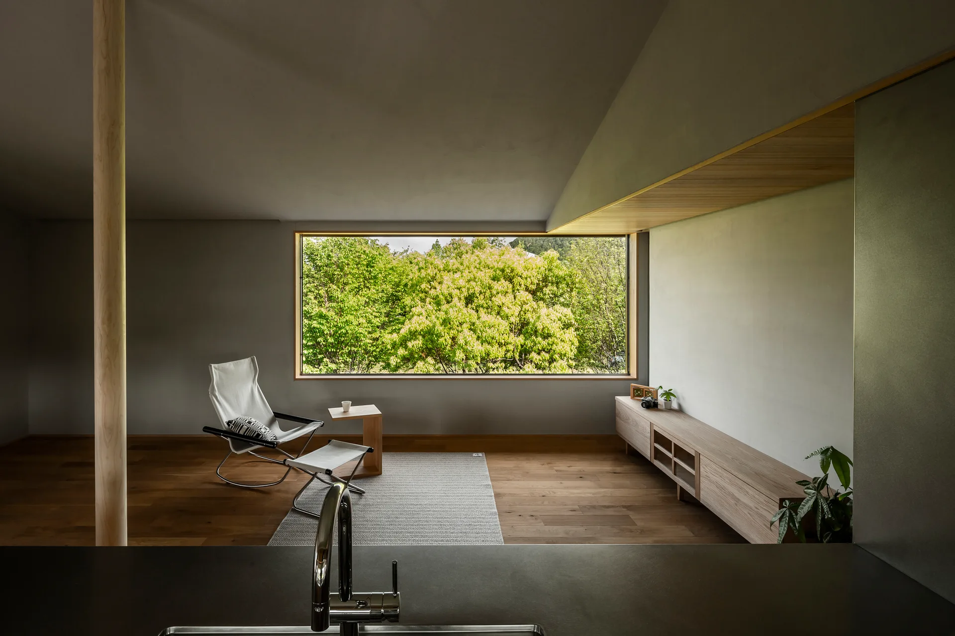 A minimalist living space features a white fabric lounge chair, a low wooden media console, and a large horizontal window showcasing lush green trees.