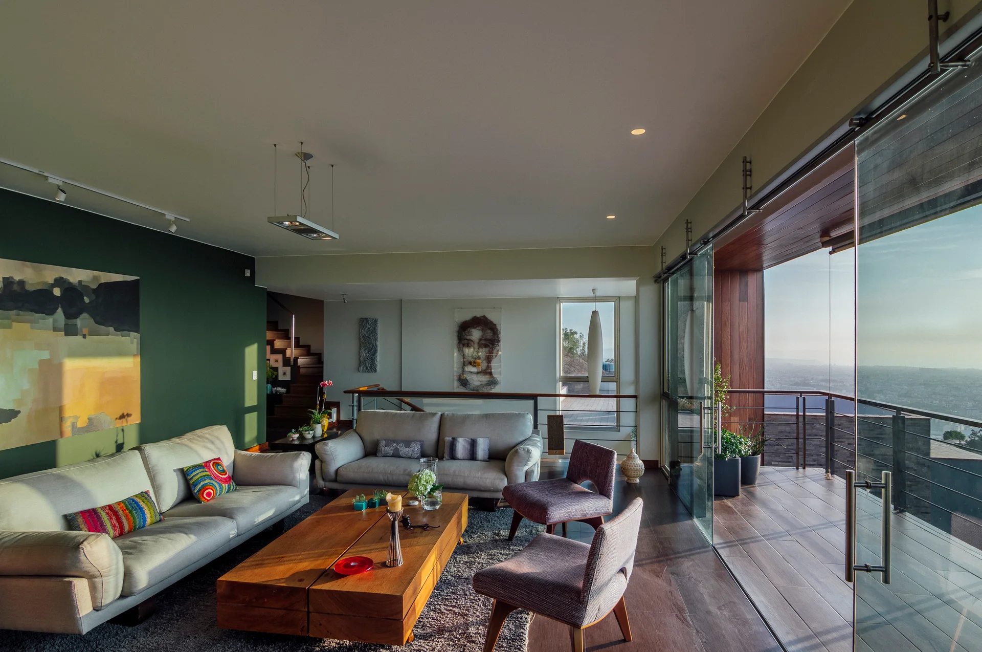 A modern living room features light gray sofas, a blocky wood coffee table, and a deep green accent wall next to sliding glass doors opening to a balcony.