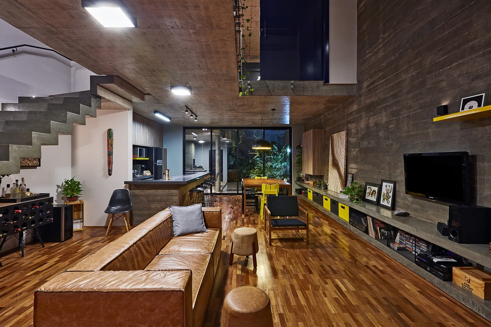 An open-concept industrial living room featuring a tan leather sofa, board-formed concrete walls, and a floating concrete staircase.