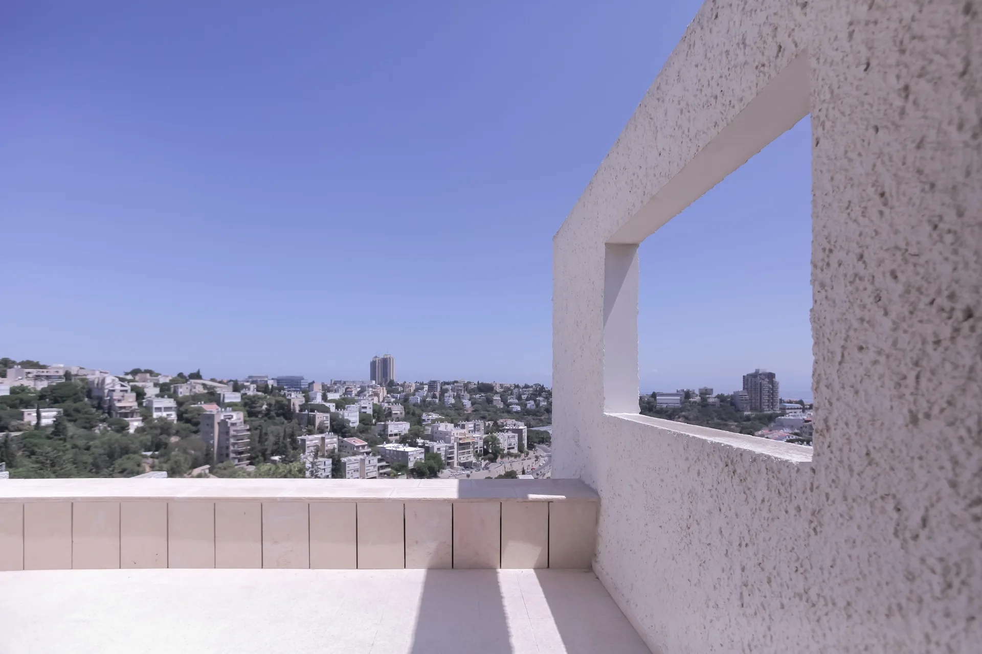 A white minimalist terrace features a textured stucco wall with a rectangular cutout framing a panoramic city view under a clear blue sky.