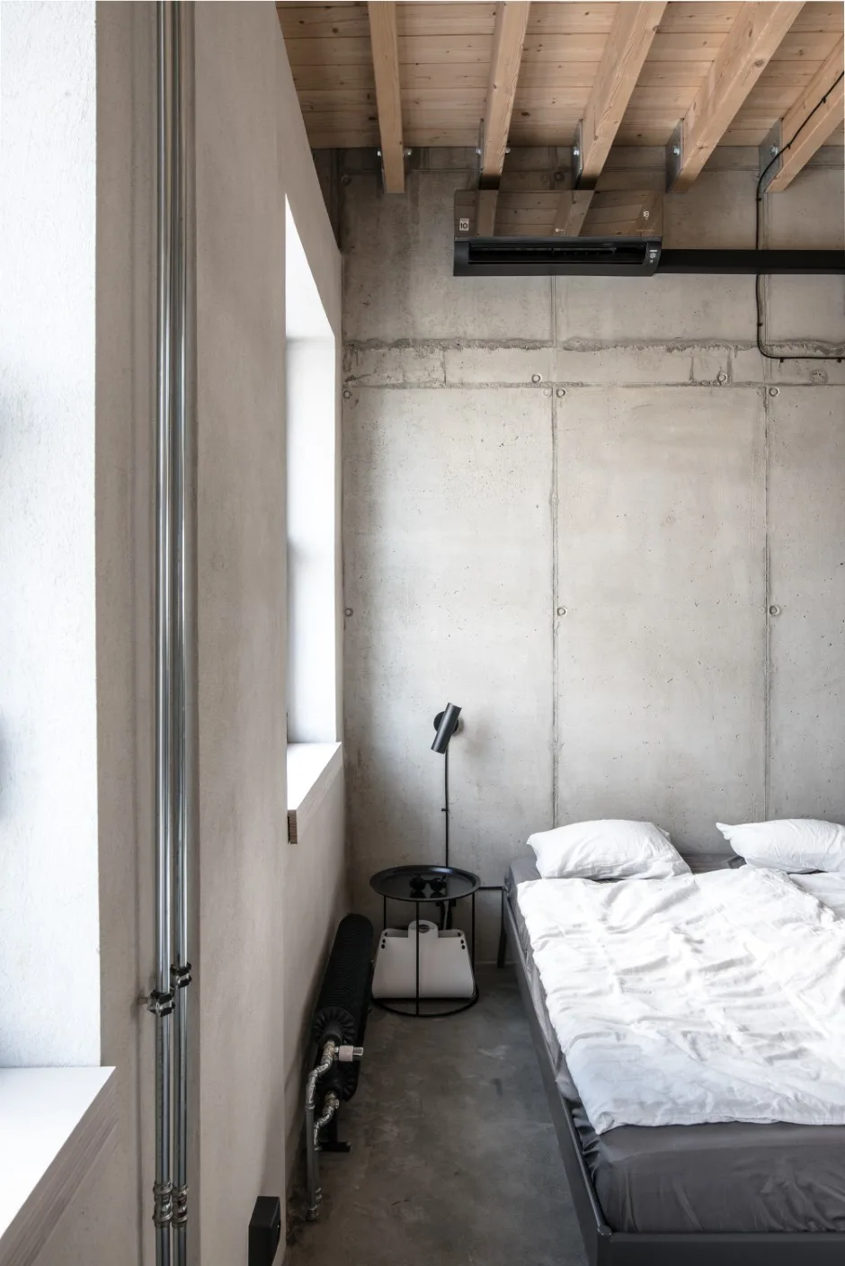 A minimalist bedroom features a bed with white sheets against a raw concrete wall beneath a ceiling with exposed natural wood joists.