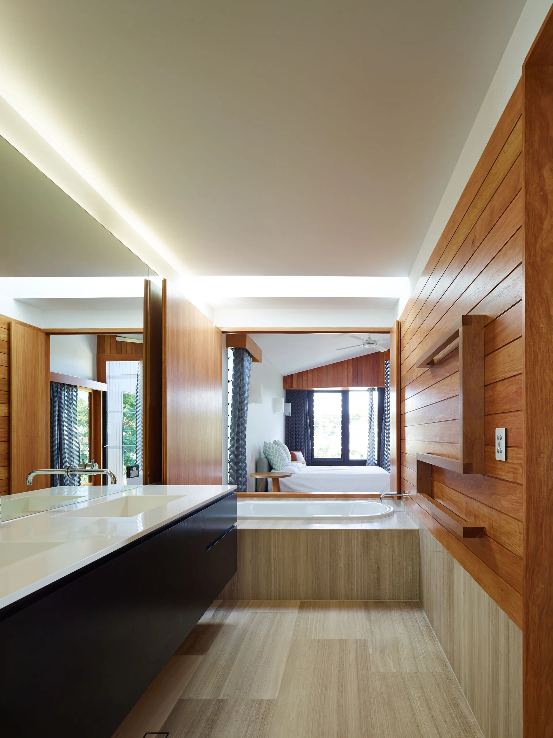 A modern bathroom features a dark floating vanity, built-in bathtub, and horizontal wood paneling, opening directly into an adjacent bedroom.