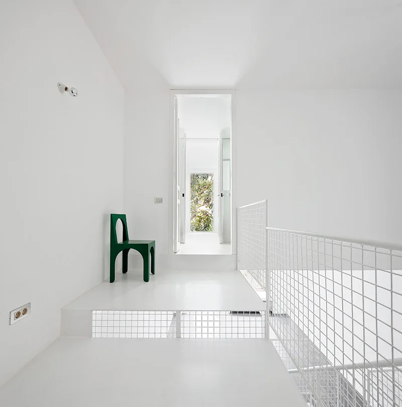 A minimalist white landing features a green geometric accent chair next to a white metal wire grid railing leading to an open doorway.