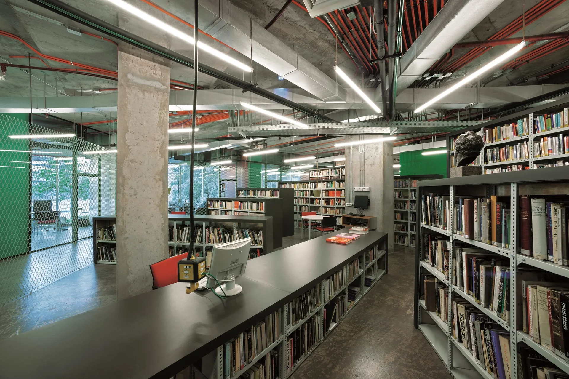 An industrial library space features rows of metal bookshelves, a long dark study table, exposed concrete pillars, and a ceiling with red conduits.