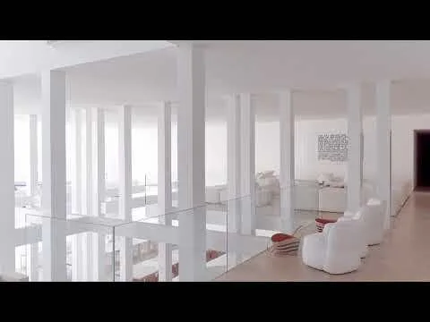 A minimalist white lounge area featuring upholstered armchairs, a grid of square architectural columns, and a transparent glass railing.