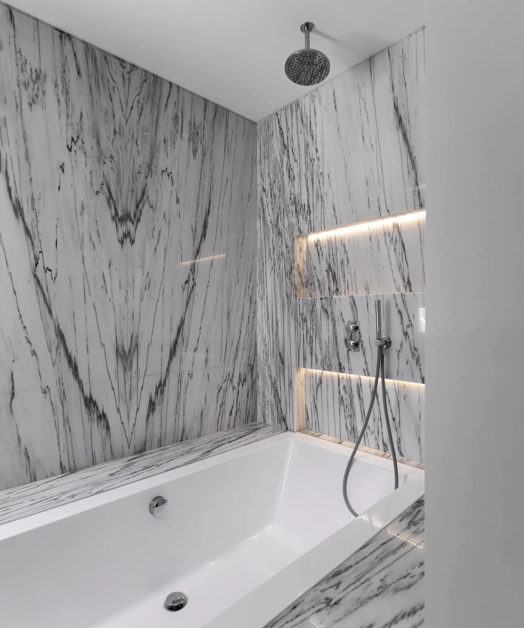 A modern white bathtub is nestled within a shower alcove clad in grey veined marble panels, featuring a recessed niche with integrated light.