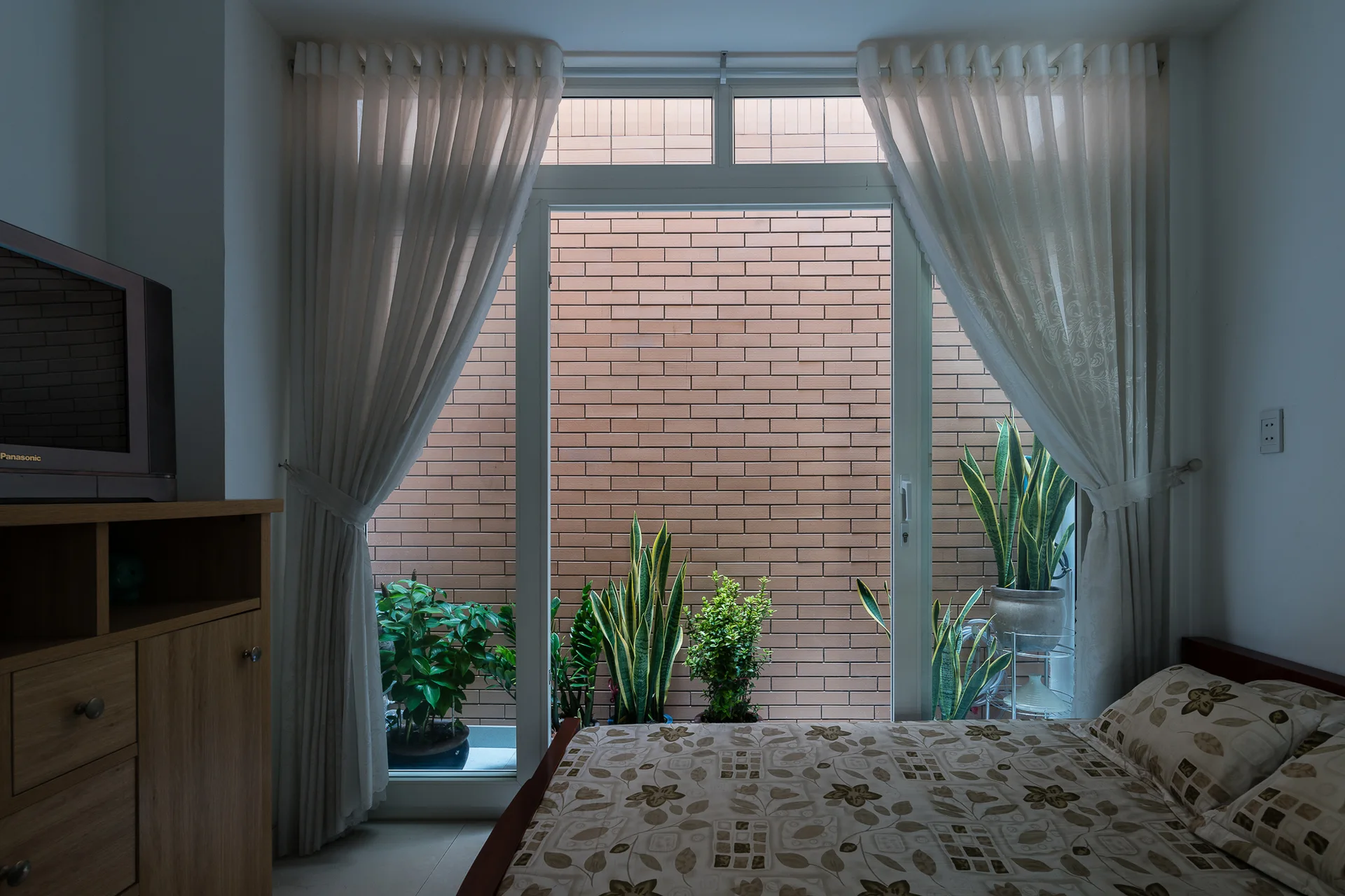 A bed with floral linens sits next to a large window with sheer white curtains opening to an outdoor brick courtyard filled with leafy potted plants.