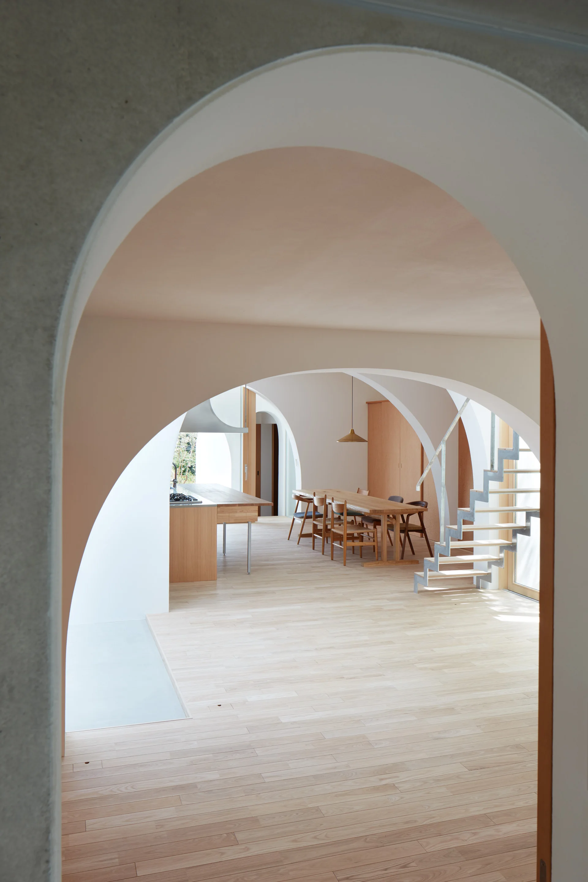 An open plan dining room viewed through a plaster archway, featuring a light wood dining table, matching chairs, and a floating metal staircase.