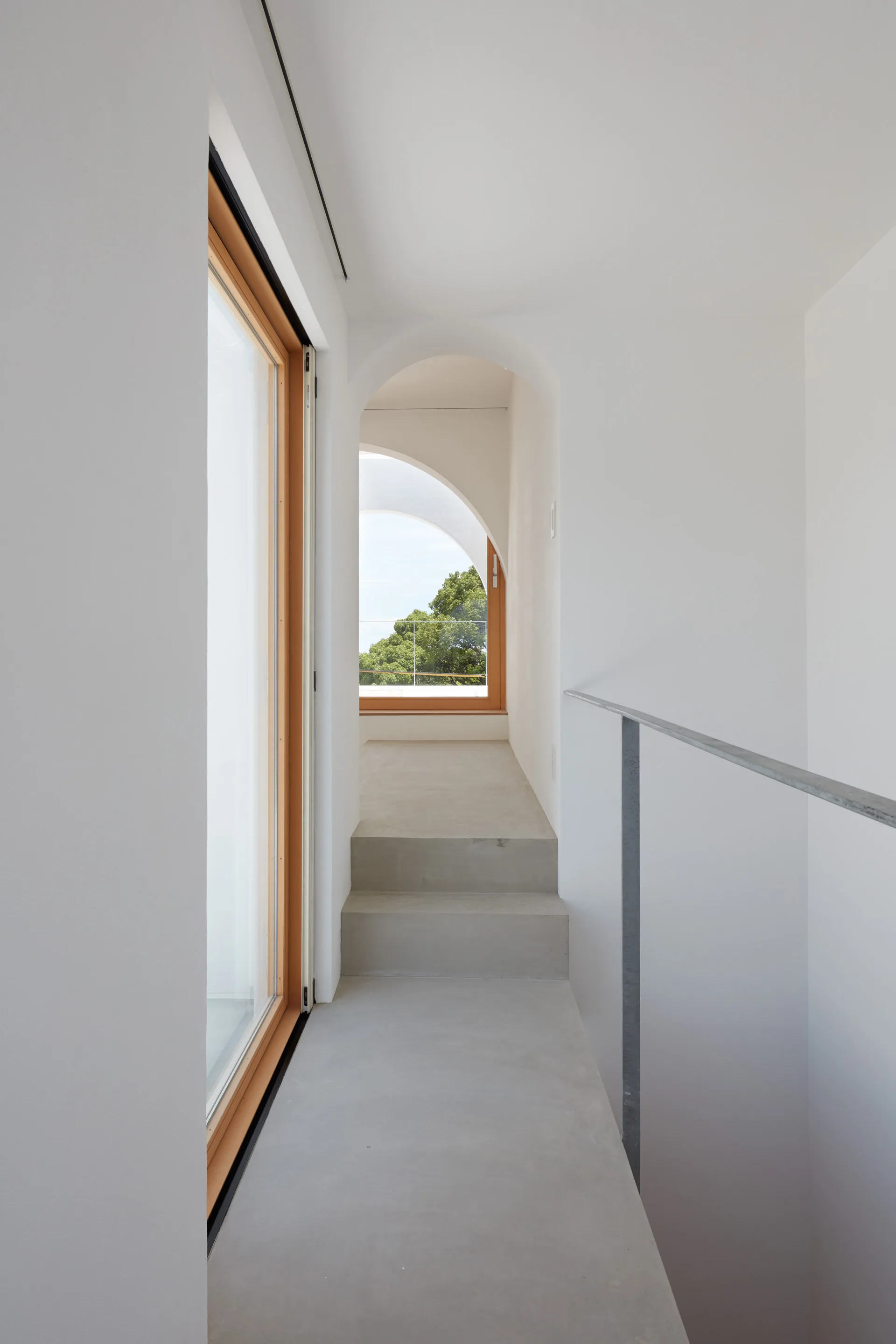 A minimalist white hallway features raw concrete steps, a slim metal handrail, and arched openings leading toward a wood-framed window.