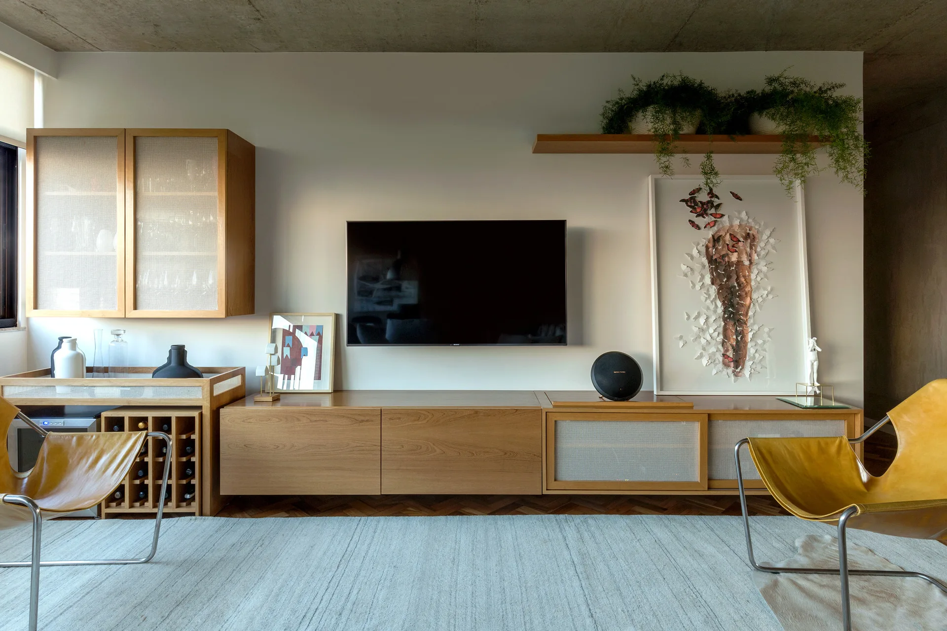 A modern living room features a long oak media console, a wall-mounted TV, two yellow leather chairs, and an exposed concrete ceiling.