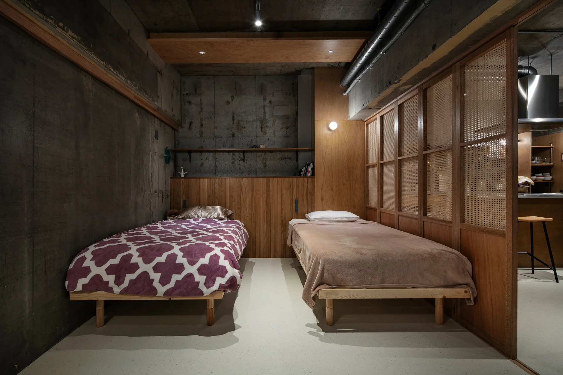 Two twin beds sit against warm wood wall paneling in a bedroom with raw concrete walls, a smooth white floor, and a cane-webbed sliding screen partition.