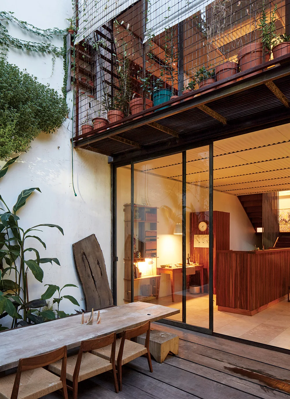 A wooden patio deck with a dining table and chairs sits next to sliding glass doors that open to a warm interior with wood cabinets and a staircase.