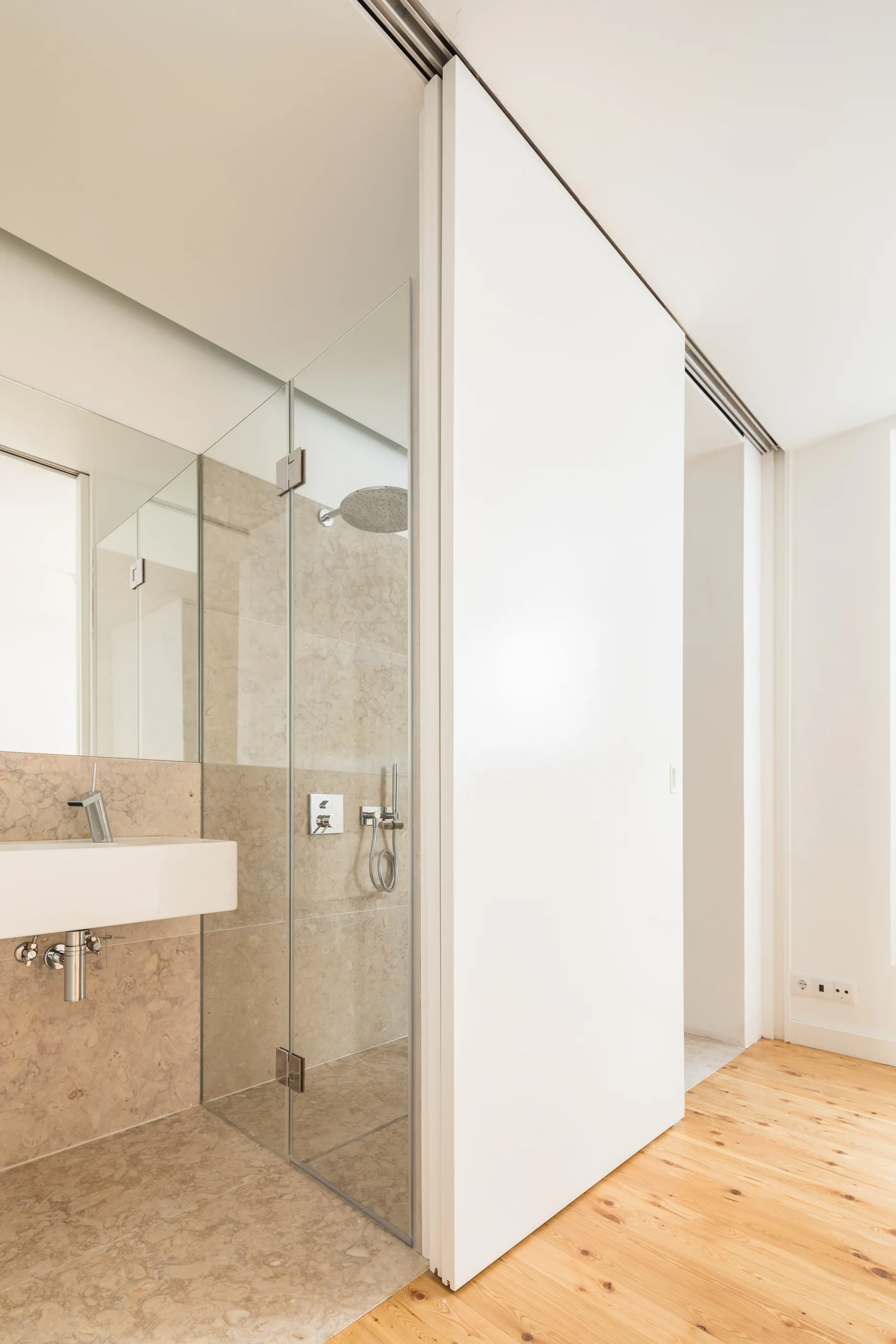 A modern bathroom features a beige stone shower enclosure next to a white wall-mounted sink and light wood flooring.