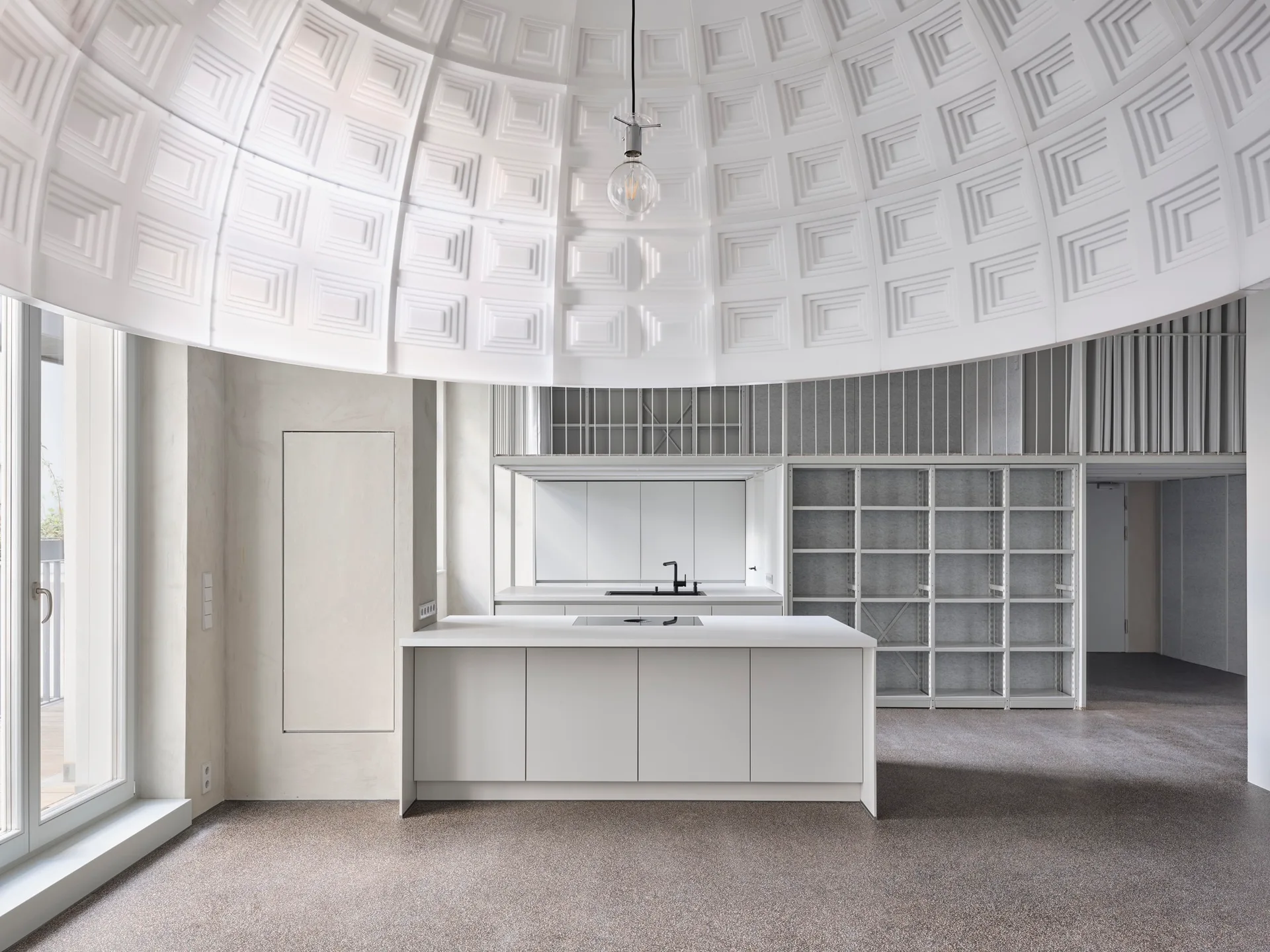 A minimalist white kitchen island stands beneath a large white coffered dome ceiling, with open metal shelving in the background.