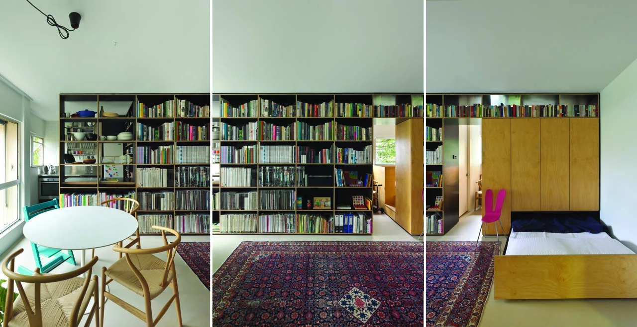 A multifunctional room features a massive plywood bookshelf wall, an integrated fold-down bed, a patterned rug, and a small dining set.