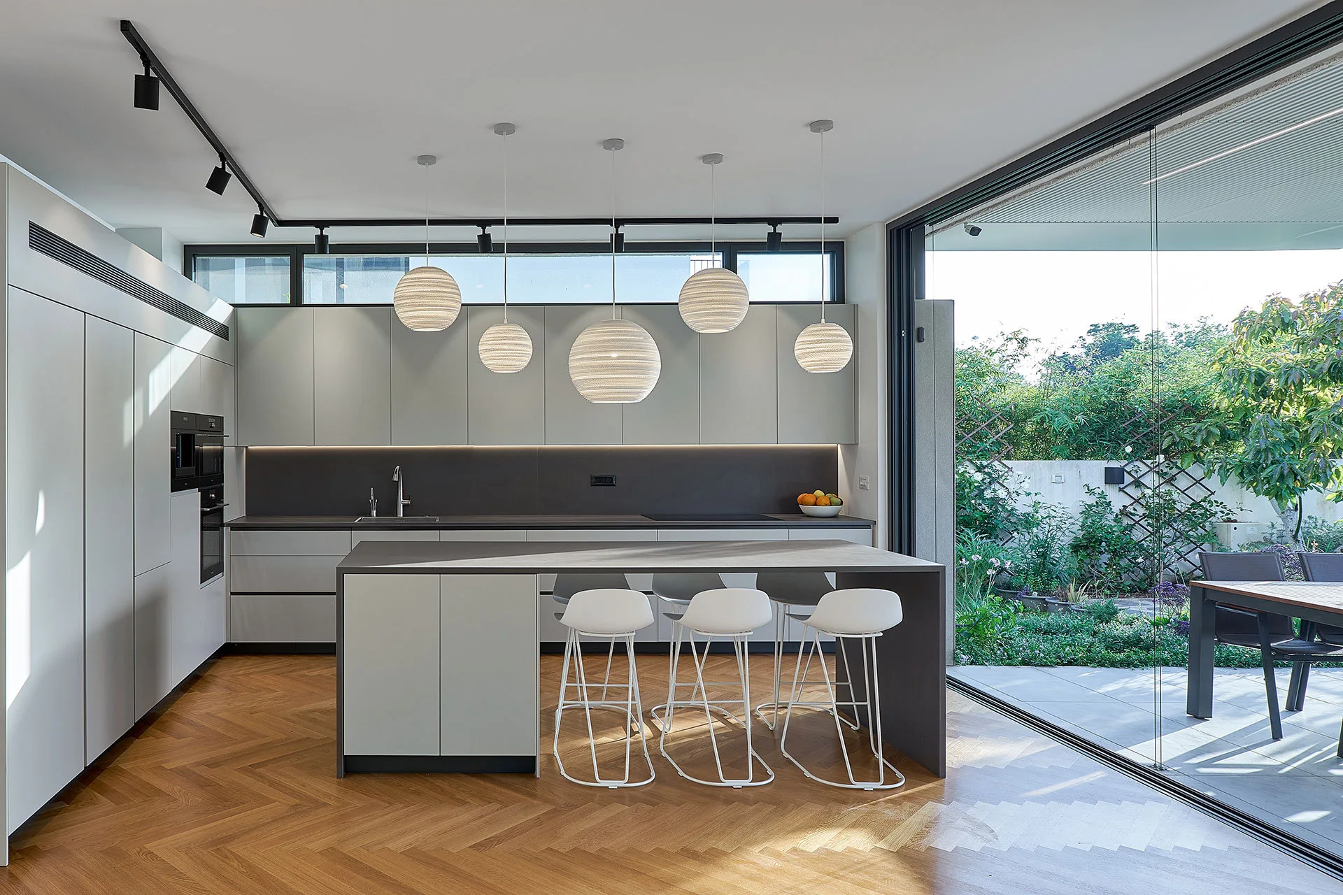 A modern grey kitchen features a central island with white bar stools, textured globe pendant lights, herringbone wood floors, and sliding glass doors.