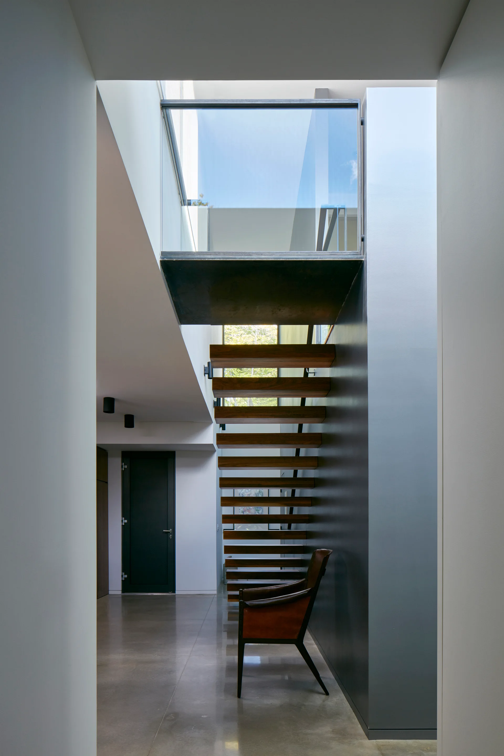 A modern hallway features a floating wooden staircase against a dark gray feature wall next to a brown leather armchair on polished concrete floors.
