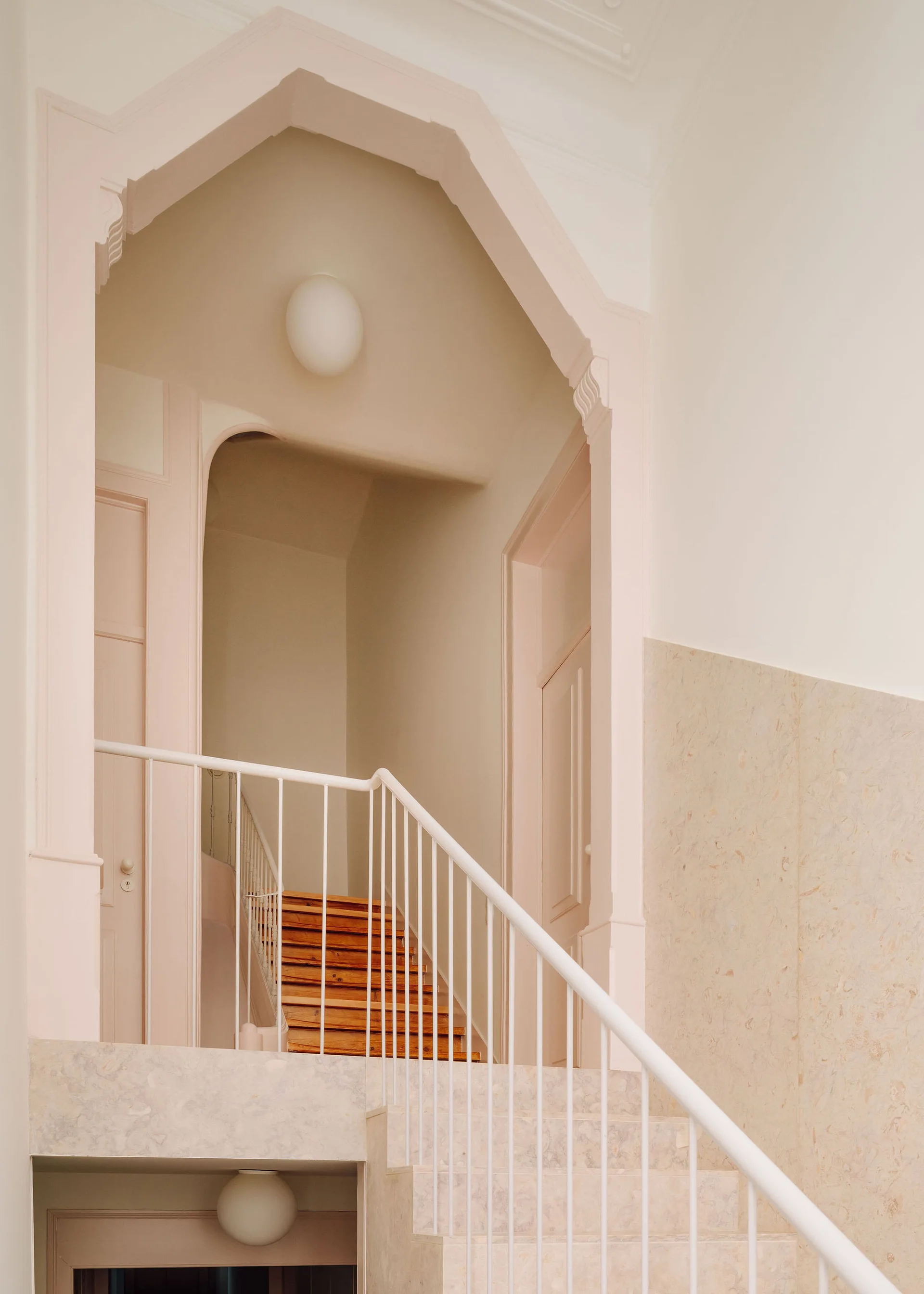 An interior view of a staircase featuring wooden steps, a white metal railing, pale pink trim, and a pointed archway.