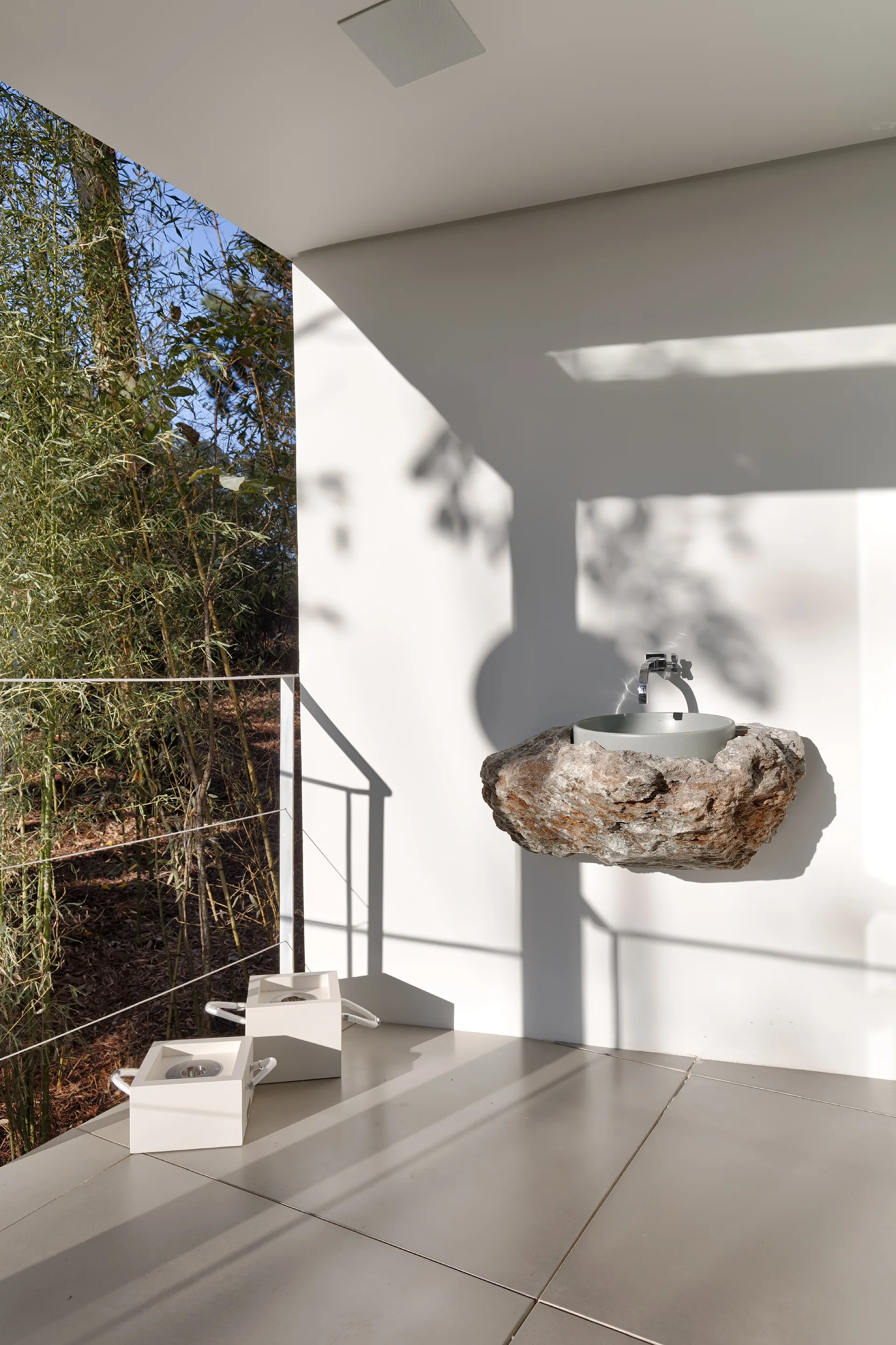 A rugged natural stone wall-mounted washbasin with a modern chrome faucet on a smooth white patio wall next to a bamboo garden.