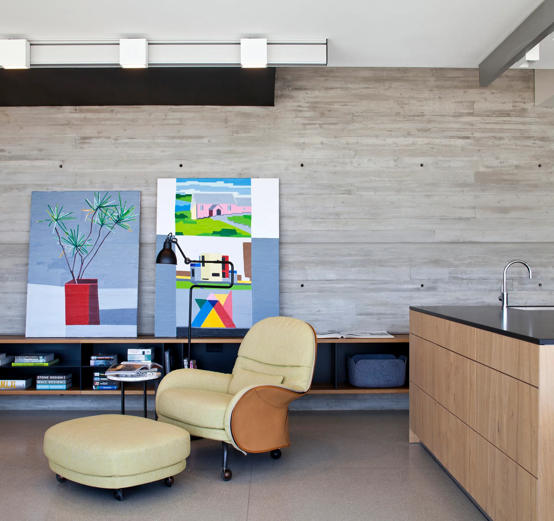 A modern living space features a yellow upholstered armchair and ottoman set against a board-formed concrete wall with leaning artwork and a low shelf.