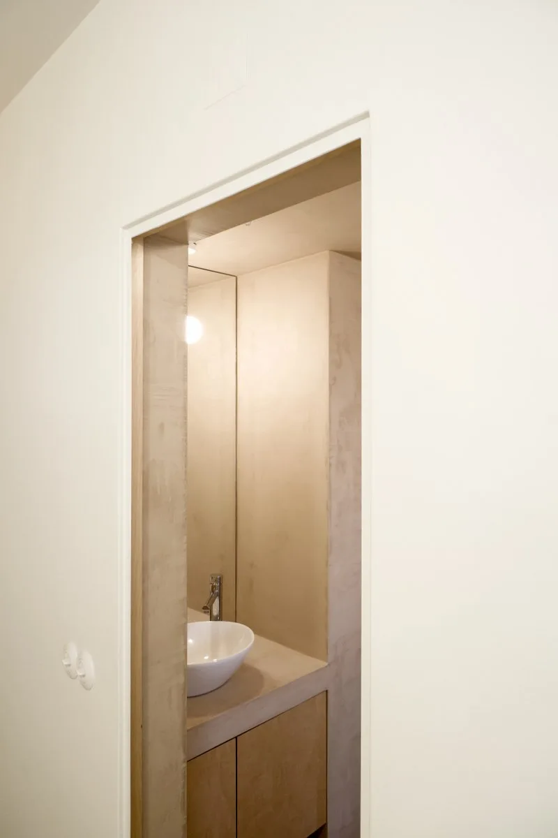 A view through a doorway into a small minimalist powder room featuring a white vessel sink on a wood vanity in a warm plaster recess.