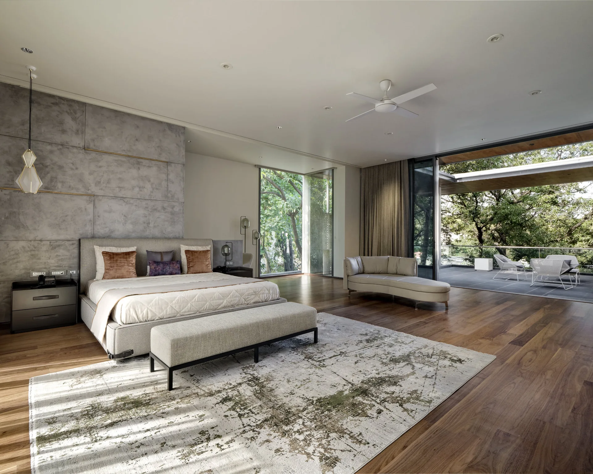 A modern primary bedroom with a grey textured accent wall, wide plank hardwood flooring, and sliding glass doors that open to a green outdoor terrace.