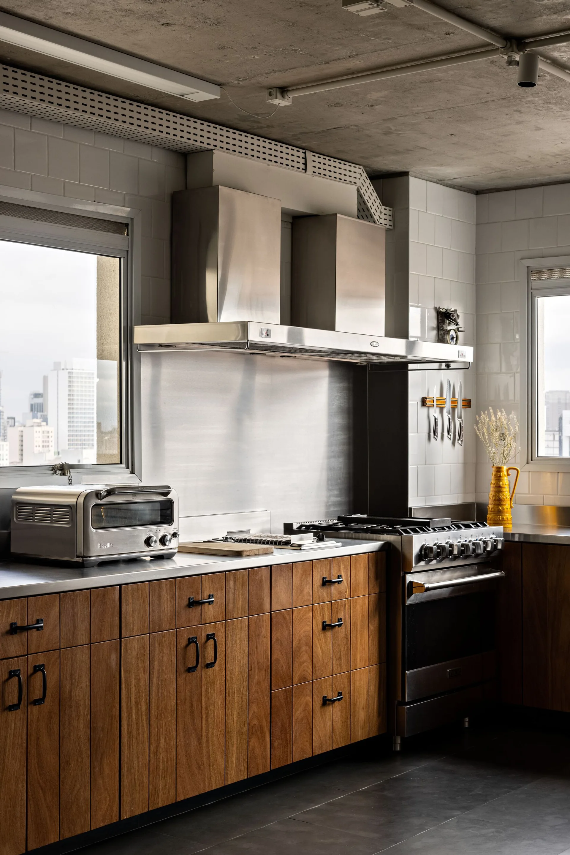 An industrial kitchen with warm wood cabinets, stainless steel countertops and appliances, white subway tile walls, and an exposed concrete ceiling.
