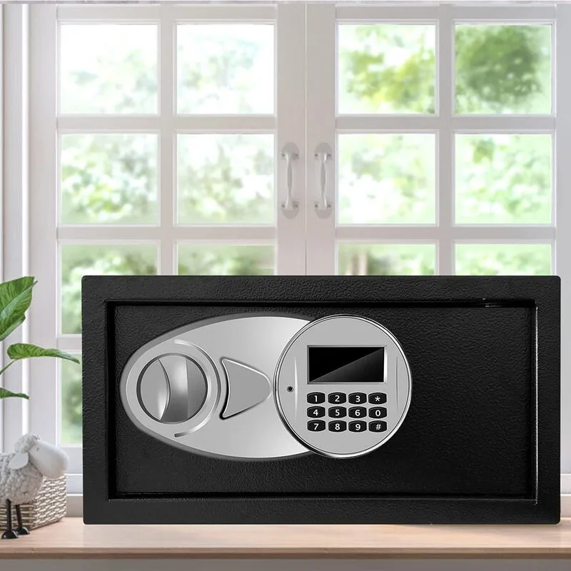 A matte black steel digital safe with a silver circular keypad sits on a light wood surface in front of a white window.