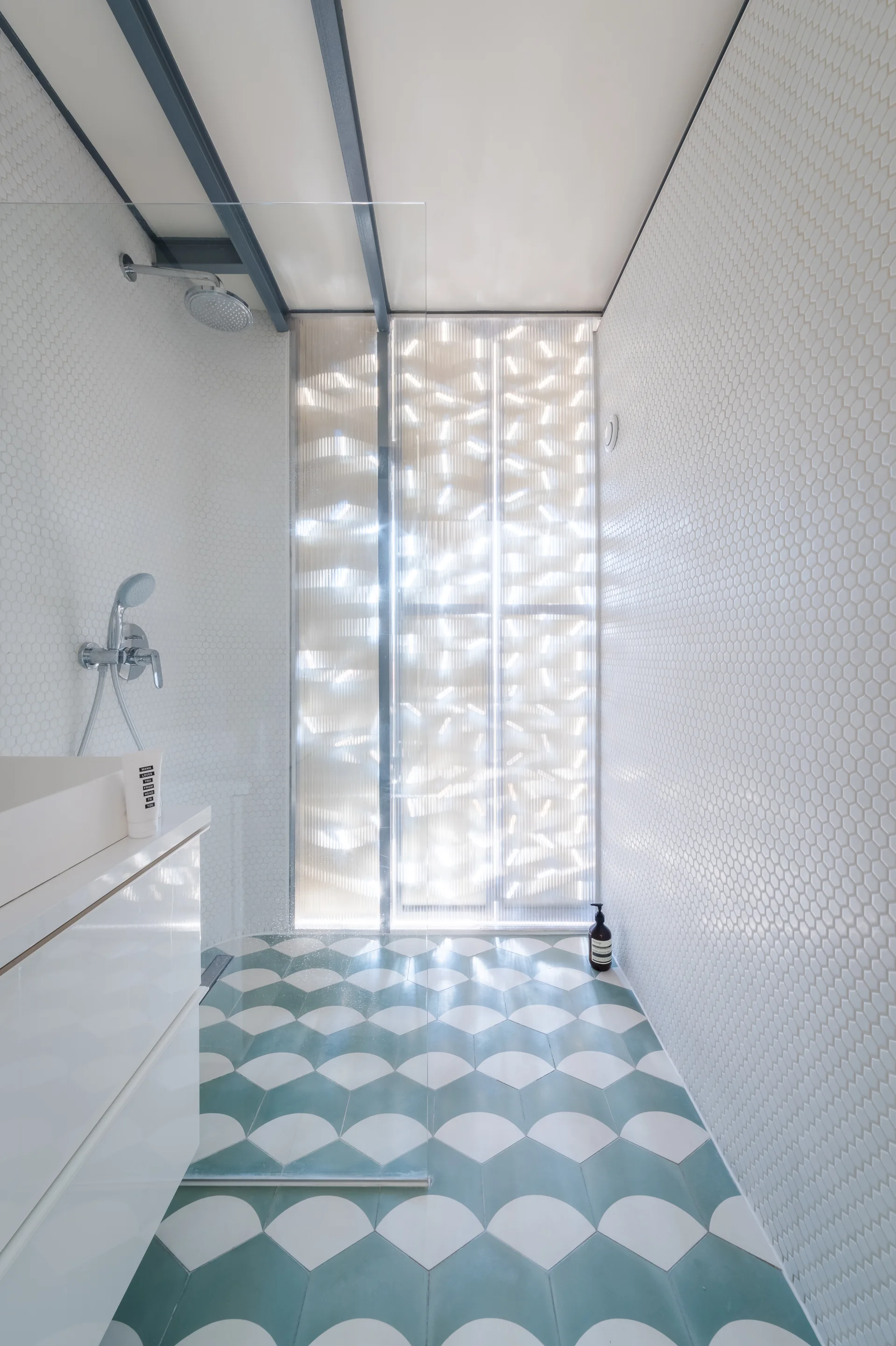 A modern walk-in shower features white hexagonal wall tiles, teal and white fan-patterned floor tiles, and a translucent fluted glass panel.