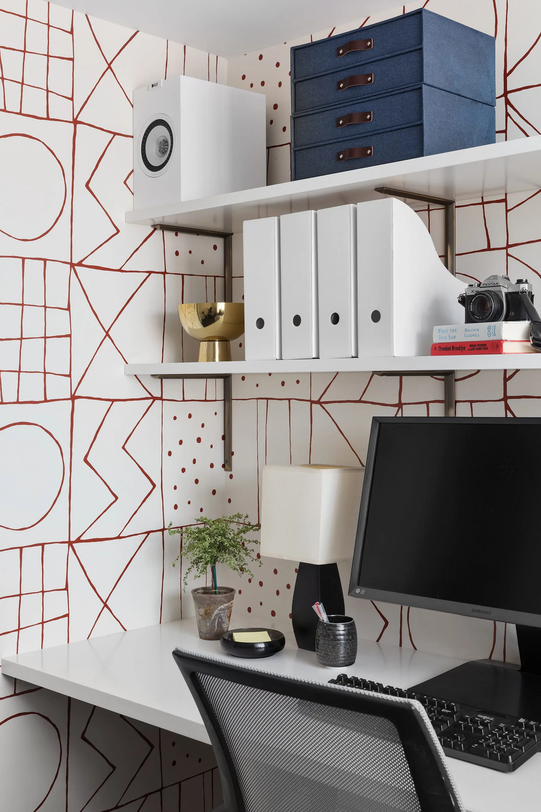 A white home office desk with a computer monitor situated below white shelves mounted on a wall with red and white geometric wallpaper.