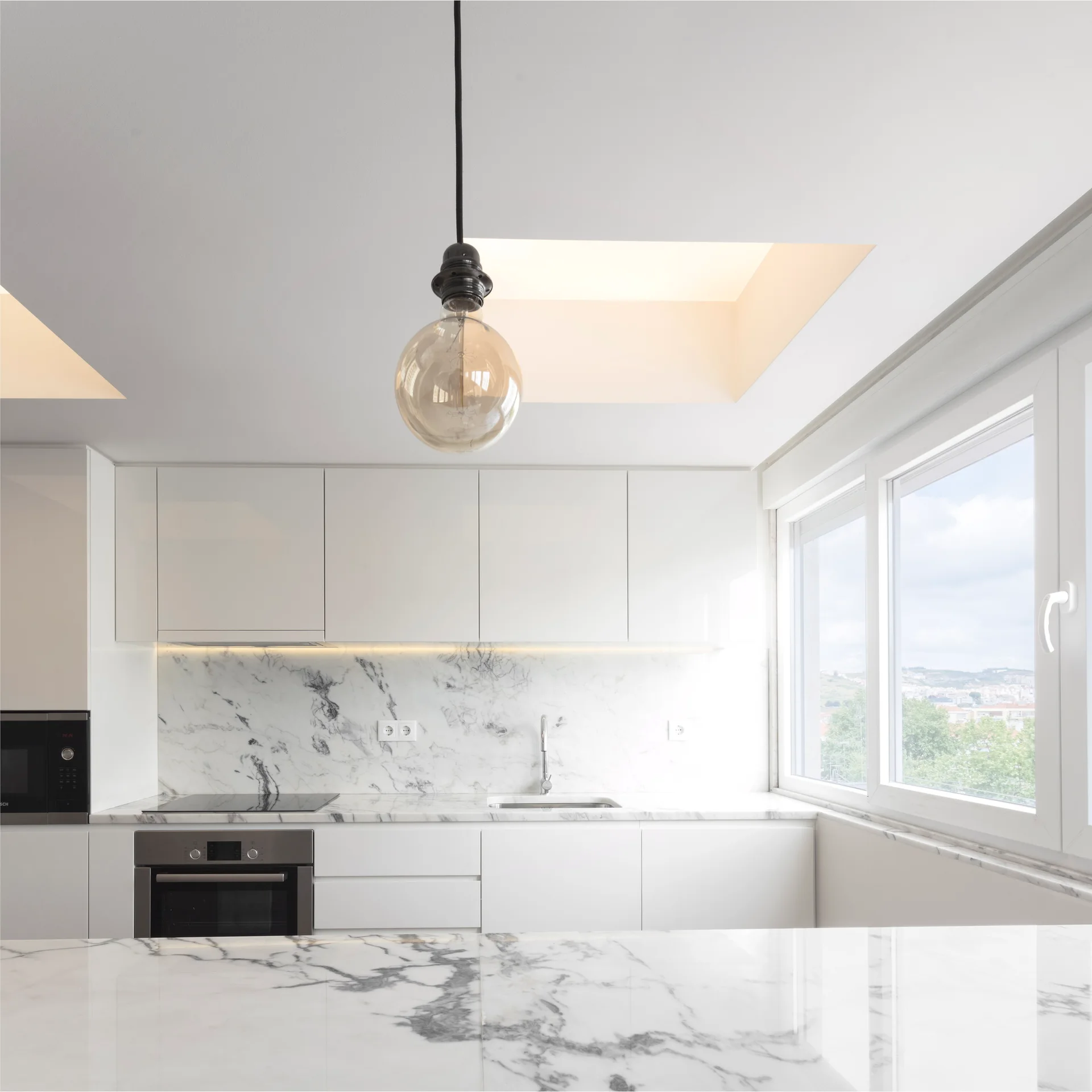 A modern white kitchen featuring a heavily veined marble backsplash, flat-front cabinetry, a built-in oven, and a glass globe pendant light.